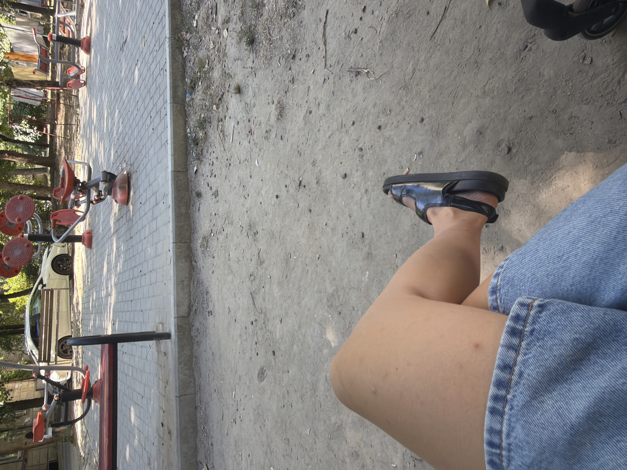 The image captures a low-angle view of a person's legs and feet, sitting on the ground in an outdoor park area. The person is wearing blue denim shorts and black sandals. The ground is dirt and gravel, with some sparse vegetation. In the background, there are several pieces of outdoor exercise equipment, a paved walkway, and a white car parked on the side of the road. Trees and foliage are visible behind the exercise equipment, suggesting a park or recreational area. The scene appears to be in Chișinău, Moldova, given the context provided.