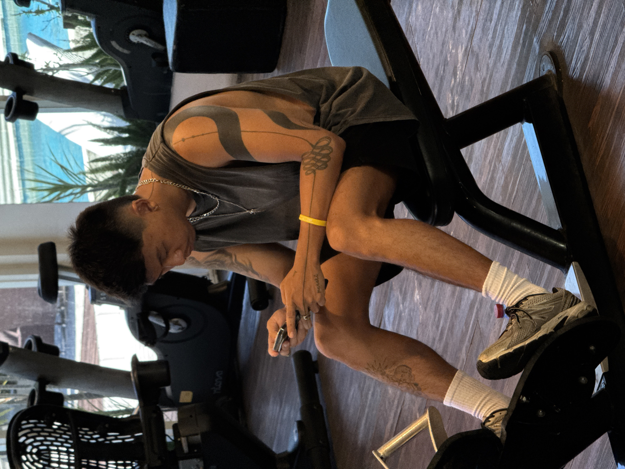 A person is sitting on a piece of exercise equipment inside what appears to be a gym. The person is wearing a grey tank top, black shorts, white socks, and athletic shoes. They have tattoos on their arms and legs and are holding an object in their hands. Gym equipment is visible in the background, and there is a window with a view of plants and possibly a body of water. The location of the image is in North Kuta, Indonesia.