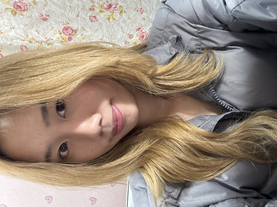 A young Asian woman with long, honey-blonde hair and fair skin lies on her side. She wears a grey puffer jacket. The background features a floral patterned blanket or wallpaper above her head. Below her head is a surface with small pink hearts on it. Her dark eyes look towards the camera with a slight smile. She wears pink lipstick.