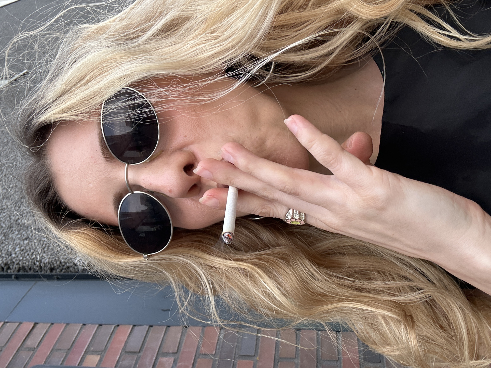 A woman with blonde hair, wearing round sunglasses and a ring on her right hand, is holding a lit cigarette near her nose. She is situated in what appears to be Amsterdam, Netherlands, as evidenced by the typical red brick paving. The background includes gray surface, black fabric and brick paving.