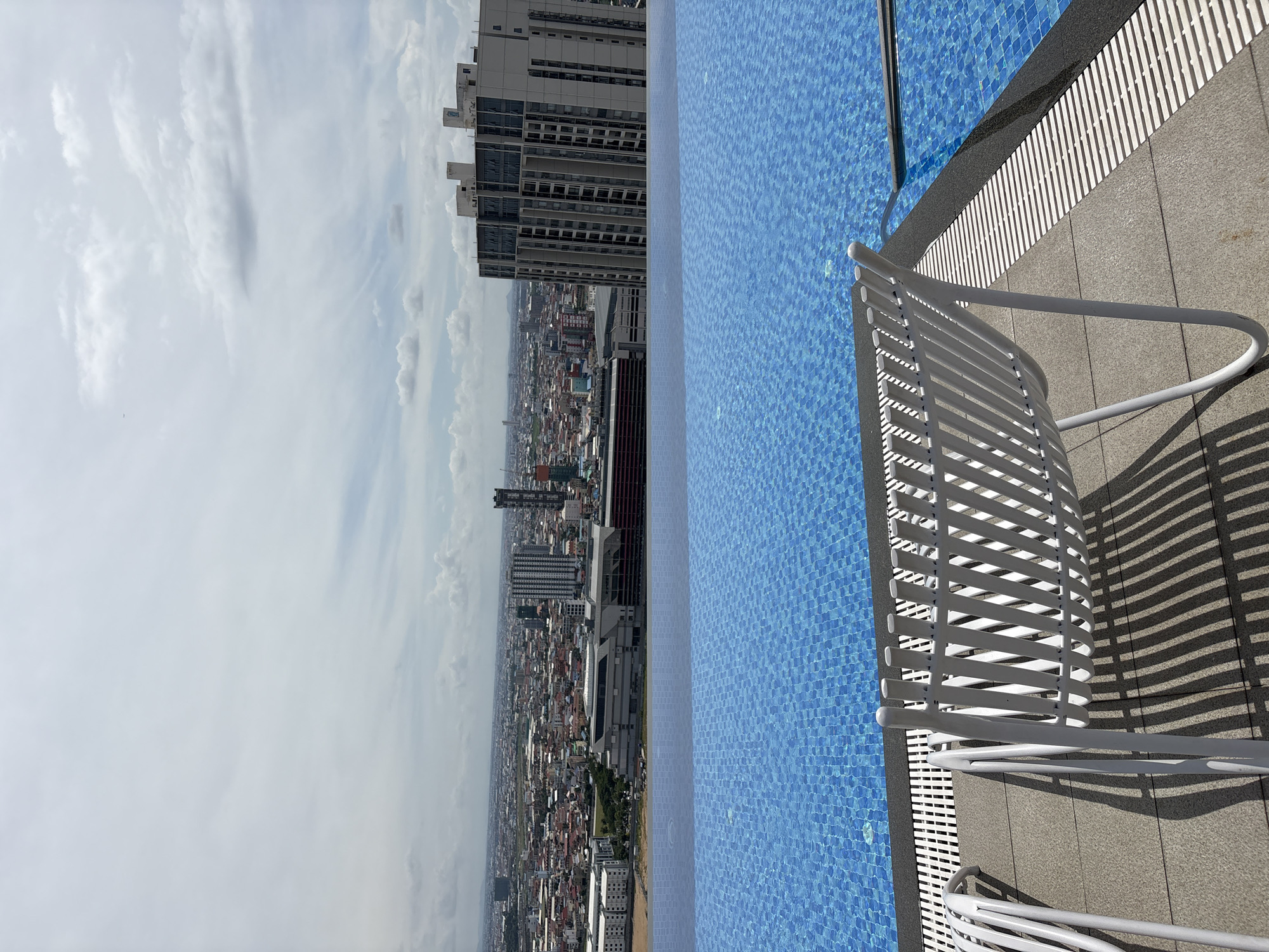 A high-angle, medium shot captures an infinity pool with a tiled blue bottom overlooking a dense urban landscape under a cloudy sky. Two white lounge chairs, made of metal with a slatted design, are positioned on the tiled deck by the pool. The pool's edge is visible, and beyond it, a vast city stretches out, featuring numerous buildings of varying heights and architectural styles, including several tall skyscrapers. The city appears to be Khan Mean Chey, Cambodia, given the context. The sky is filled with white and gray clouds, suggesting a partly overcast day. The scene is devoid of people, focusing solely on the architectural elements and the urban vista.