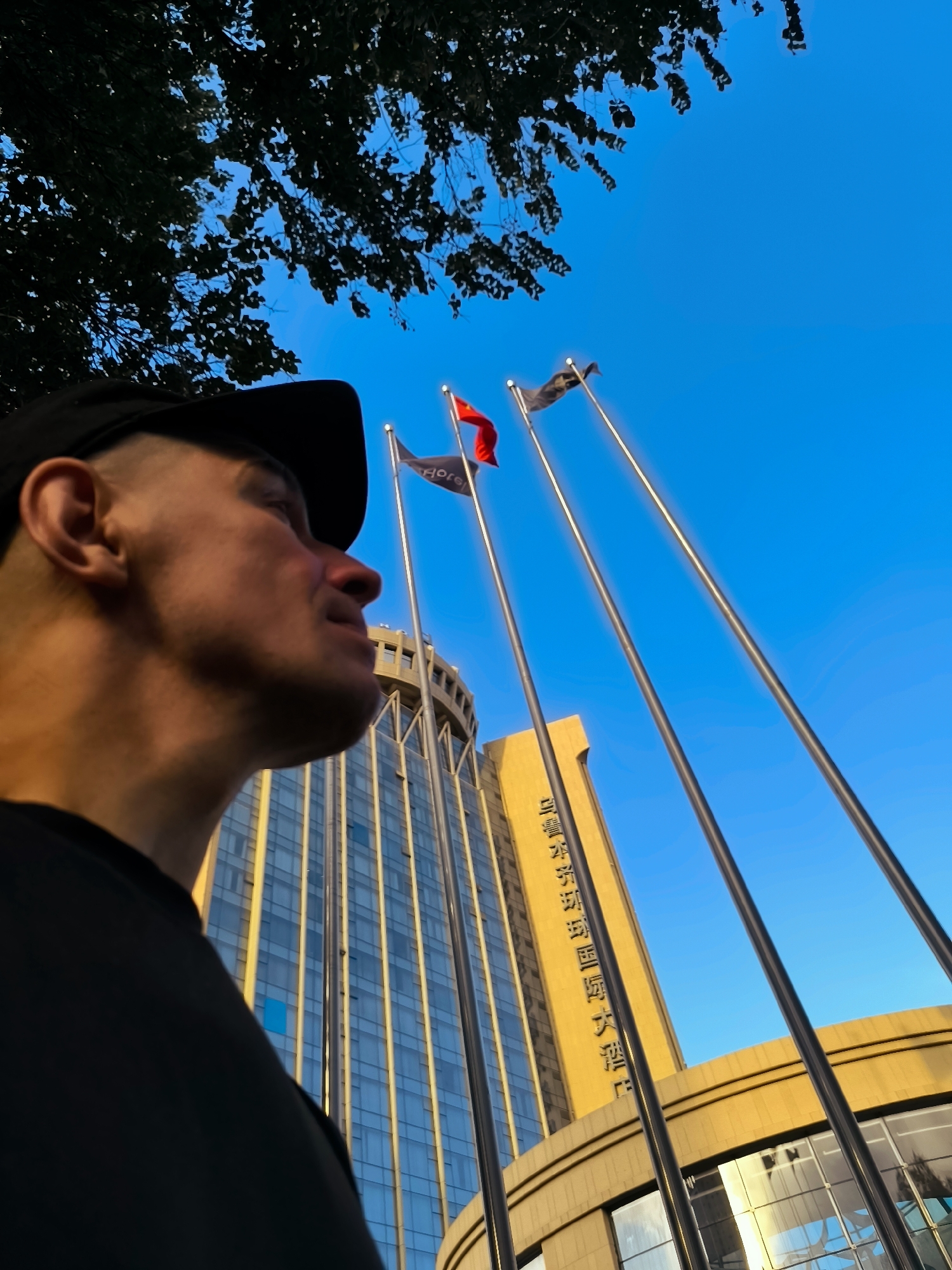 A low-angle medium shot captures a man wearing a black t-shirt and a black baseball cap in profile, facing towards the right side of the frame. The man's skin is illuminated by warm, golden light, highlighting his features and the side of his neck. Behind him, a tall, modern building with a yellow facade and reflective blue glass windows rises into a clear, bright blue sky. The building features vertical white accents, and large, dark vertical poles or flagpoles stand in front of it. One flag, colored red, is visible on a pole on the left, while a dark flag is visible on another pole further to the right. Tree branches, silhouetted against the sky, are visible in the upper left corner of the frame. The building appears to be the Grand Hotel in Ürümqi, China, given the Chinese characters visible on its facade.