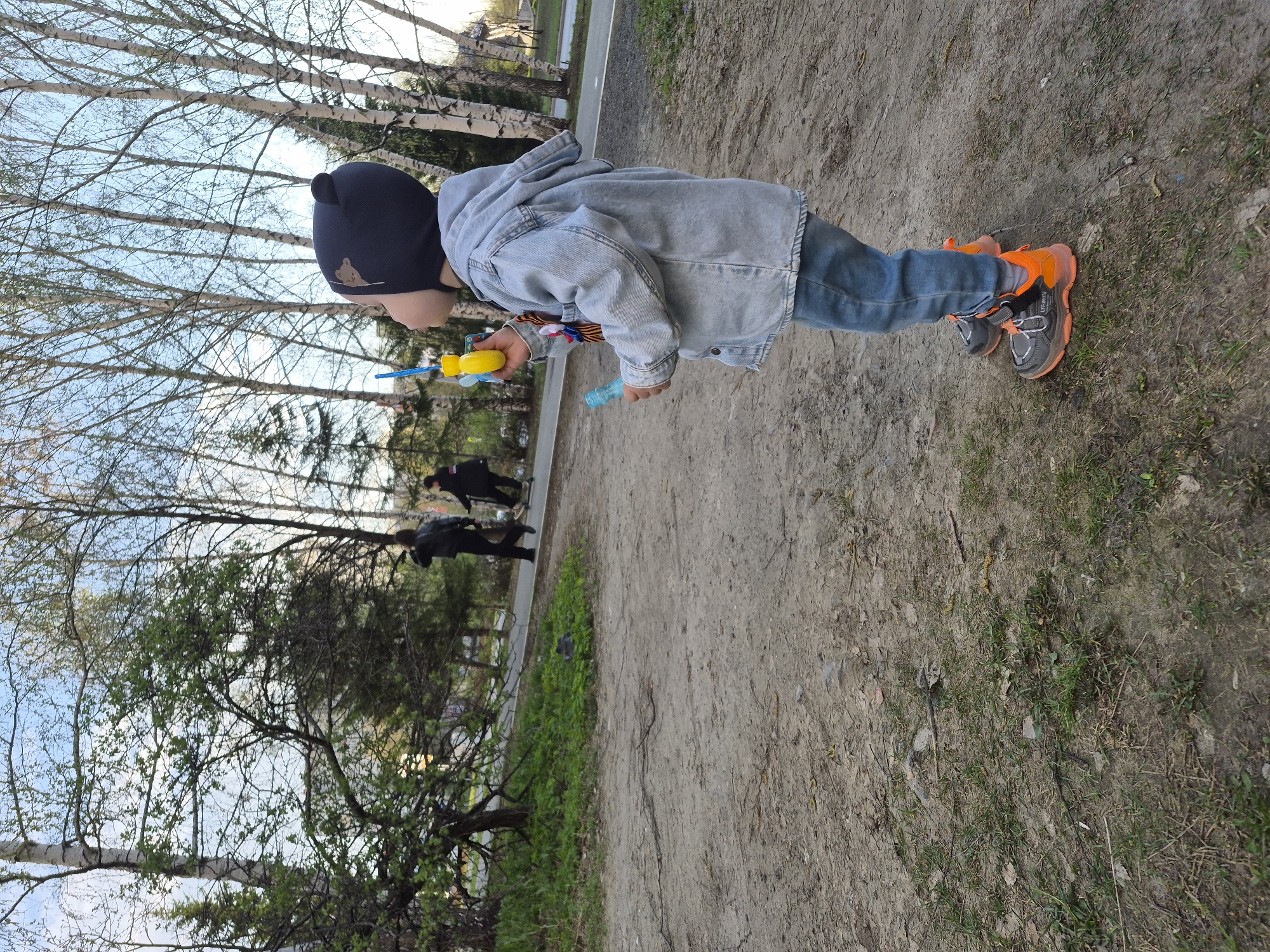 A young child stands on a dirt path in a park, seemingly in Tobolsk, Russia, based on visual cues and common imagery. The child wears a black beanie, a light denim jacket, blue jeans, and gray and orange athletic shoes. The child is holding a yellow and blue bubble wand and a blue bottle of bubble solution. A St. George ribbon is attached to the child's jacket sleeve. Two adults walk in the background. Trees and a partial fence border the path. The ground is a mix of dirt and sparse grass. Bare branches indicate the beginning of spring.
