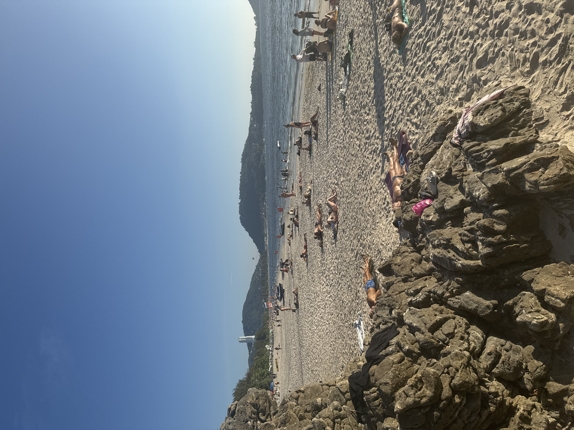 This image depicts a beach scene in Patong, Thailand. The scene is captured from a high vantage point, likely on a rocky outcrop. The foreground consists of large, dark-colored rocks. The beach extends into the midground, populated by numerous people sunbathing, walking, and engaging in water activities near the shore. The ocean, stretching to the horizon, appears calm. In the background, there is a landmass, possibly a headland or island. The sky is clear and blue.