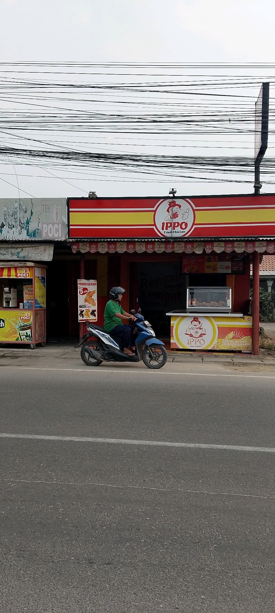 A man wearing a green shirt and dark pants rides a blue motorcycle on a paved road. He wears a black helmet. The man is on the right side of the frame. Behind him, a storefront with a red and yellow awning displays "IPPO Fried Chicken" in red lettering with a cartoon pig graphic. To the left of the storefront, another stall displays "TEH POCI". A display case in front of the "IPPO" store contains fried chicken. Power lines are strung across the top of the frame. The sky is overcast and white. The scene appears to be in Sriamur, Indonesia.