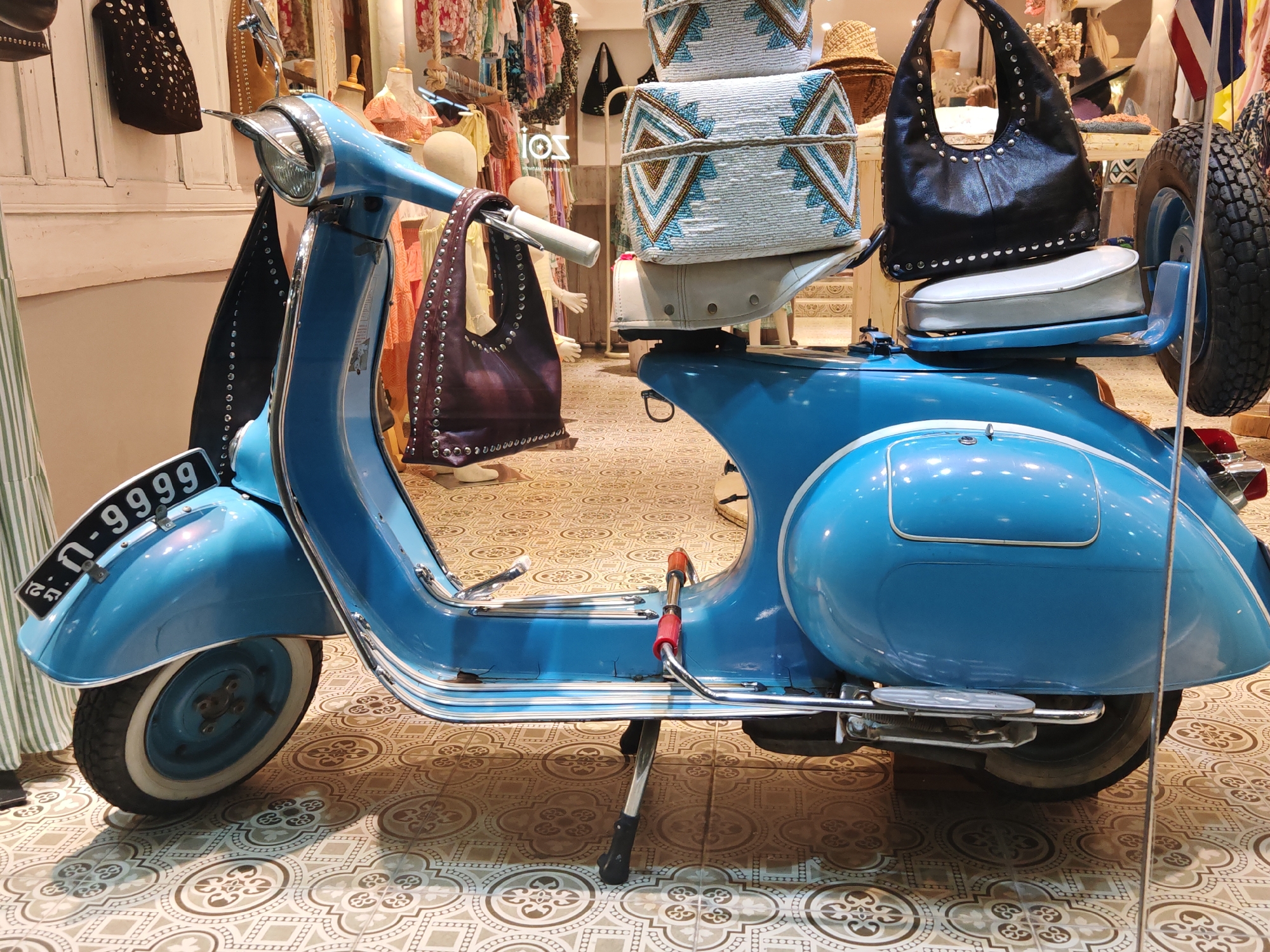 This is a medium shot, eye-level photograph of a light blue vintage Vespa scooter parked indoors in front of a retail display. The scooter is positioned diagonally, with its front on the left and rear on the right, facing towards the viewer's right. The scooter has chrome accents, a white seat, and a spare tire mounted on the rear. The license plate reads "ก-9999". Behind the scooter, various bags and clothing items are displayed on mannequins and shelves. A woman in a green and white striped garment is partially visible on the far left. The background suggests a boutique or shop with patterned floor tiles. The scene is well-lit, with no discernible shadows. The location is identified as Baan Bo Phut, Thailand.