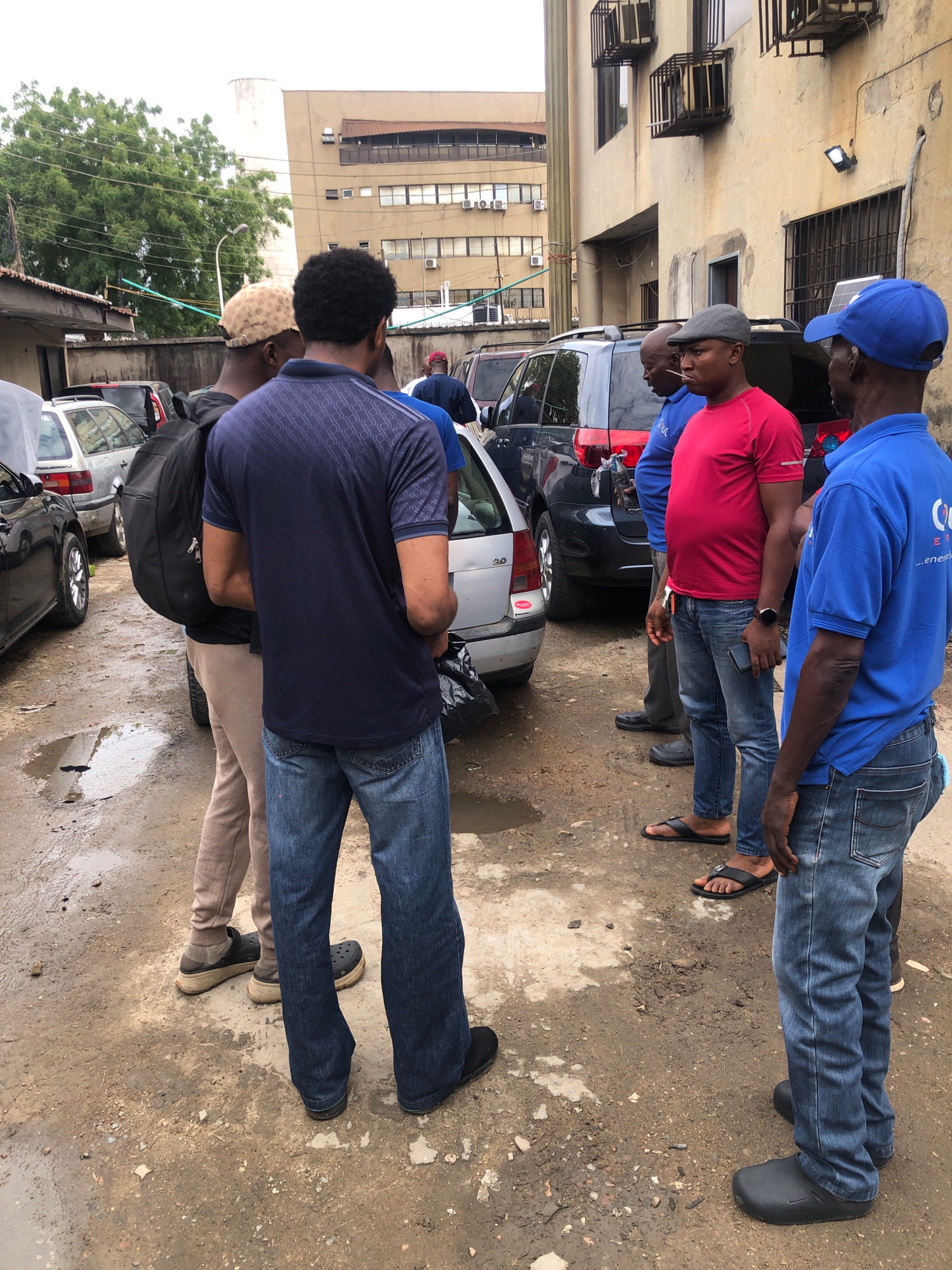 A group of five men are standing in a lot on a cloudy day in Ikeja, Nigeria, with several cars parked around them. The ground is dirt and partially covered in puddles of water, indicating recent rain. The men are dressed in casual clothing. The man closest to the viewer on the right is wearing a blue t-shirt and blue jeans. To his left is a man in a red t-shirt and blue jeans. To his left, a man in a blue t-shirt and blue jeans is facing away from the camera. Further left, a man in a patterned navy blue shirt and blue jeans is also facing away from the camera, with a black backpack on. On the far left, a man wearing a tan shirt and light-colored pants is partially visible. Two of the men are wearing hats, one a beige patterned cap, the other a grey flat cap. The environment appears to be an urban setting, with buildings visible in the background.