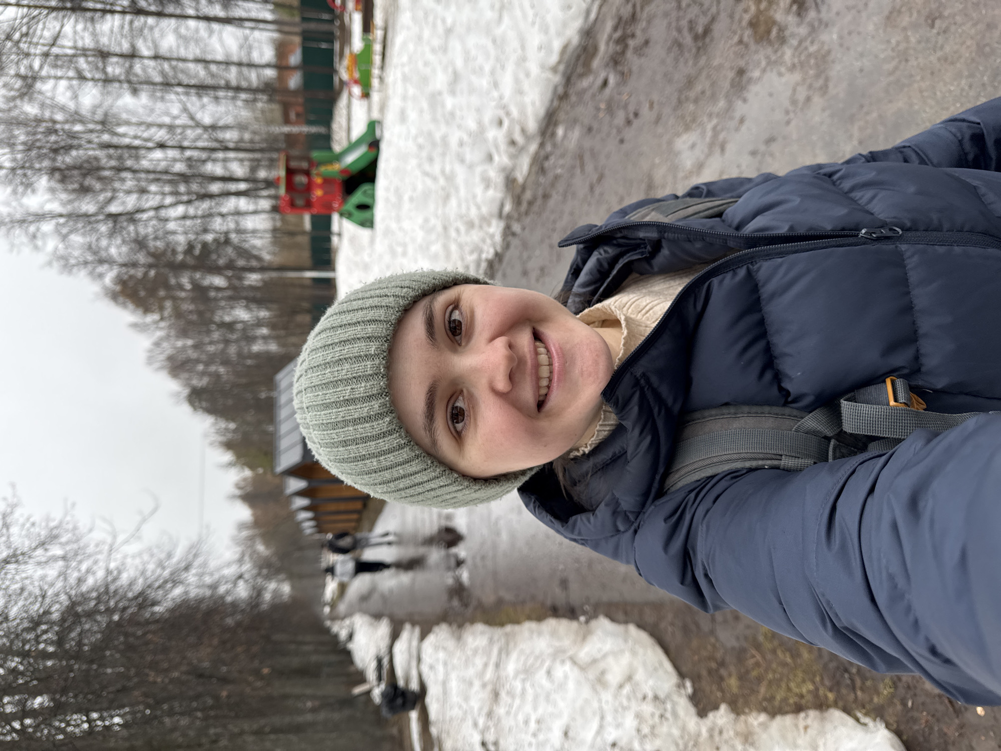 A young woman is taking a selfie outdoors in Izhevsk, Russia, with a light green beanie and a dark blue puffer jacket. She is smiling at the camera. There is patchy snow on the ground. In the background, there are trees and a children's playground with red, green, and yellow equipment. Also visible are what appear to be stalls and some blurred people in the distance. The sky is overcast.