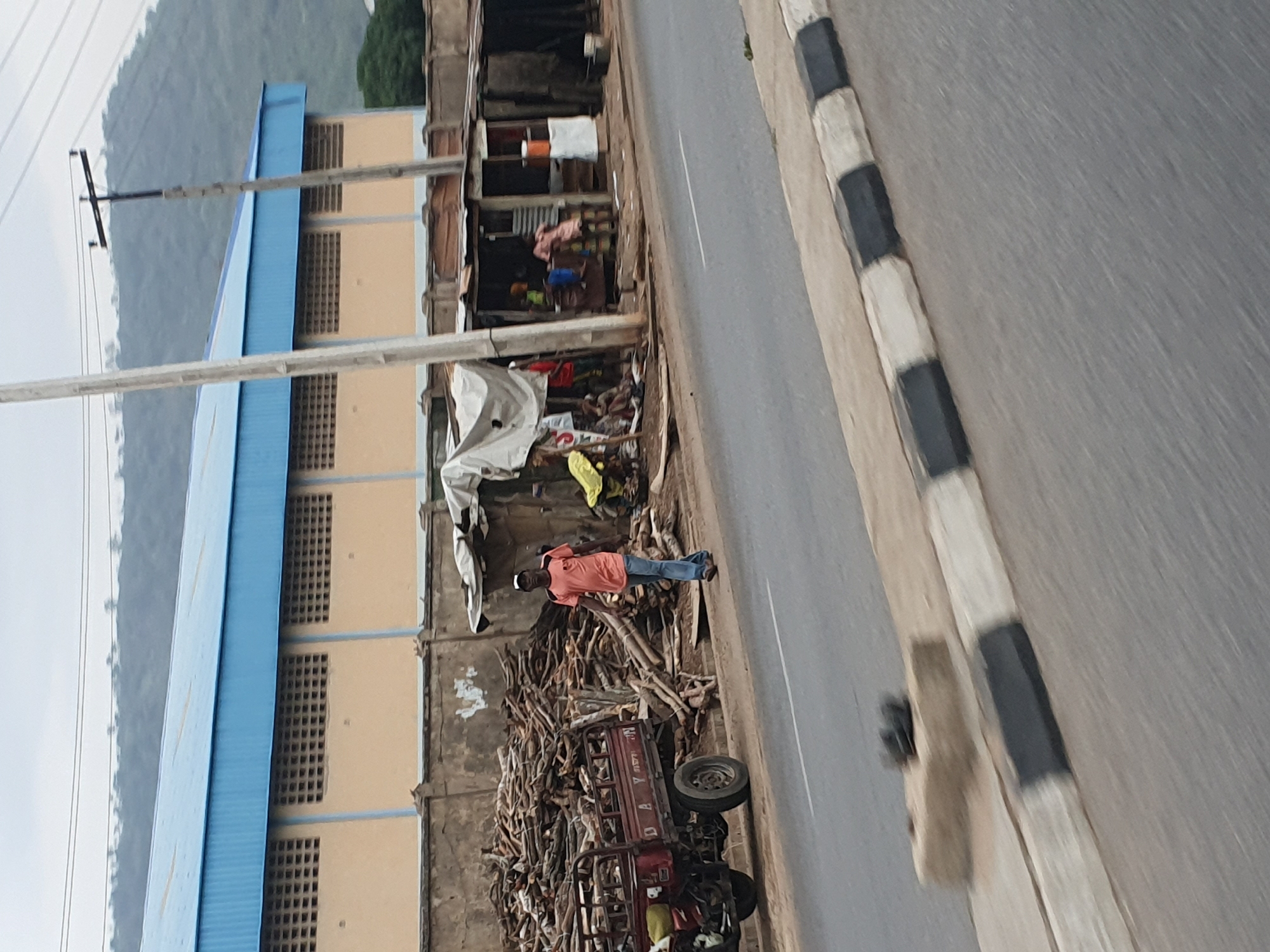 A slightly angled, medium-shot photograph shows a roadside scene in Lokoja, Nigeria. The main subject is a man in a coral-colored shirt and blue jeans, standing beside a large pile of firewood. The firewood is adjacent to a short, beige wall with an attached building that has a blue roof. A small, three-wheeled vehicle is partially obscured beneath the wood pile.
To the right of the wood pile is a gray asphalt road with a painted curb featuring alternating black and white stripes. On the left side of the road, there is a two-story building.
Behind the shop, a mountainous region is visible in the background. Overhead electrical wires and utility poles are also visible. The overall environment suggests a low-income commercial or residential area.