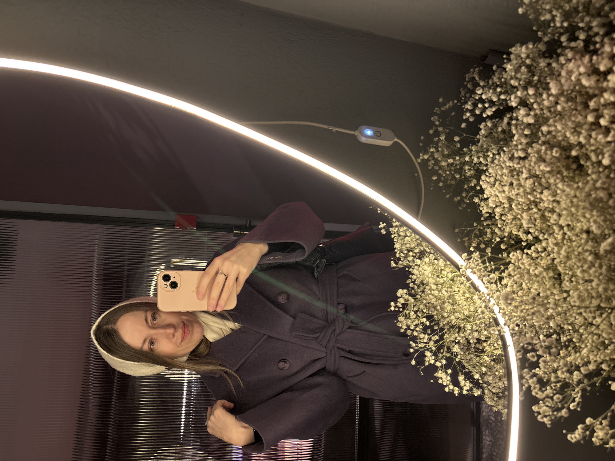 A woman is taking a mirror selfie in an enclosed space decorated with a halo light and baby's breath. She is wearing a dark purple coat with a tie belt and a cream-colored head covering. The background includes a textured wall and a dark-colored wall above. It is likely in the city of Saint Petersburg, Russia.