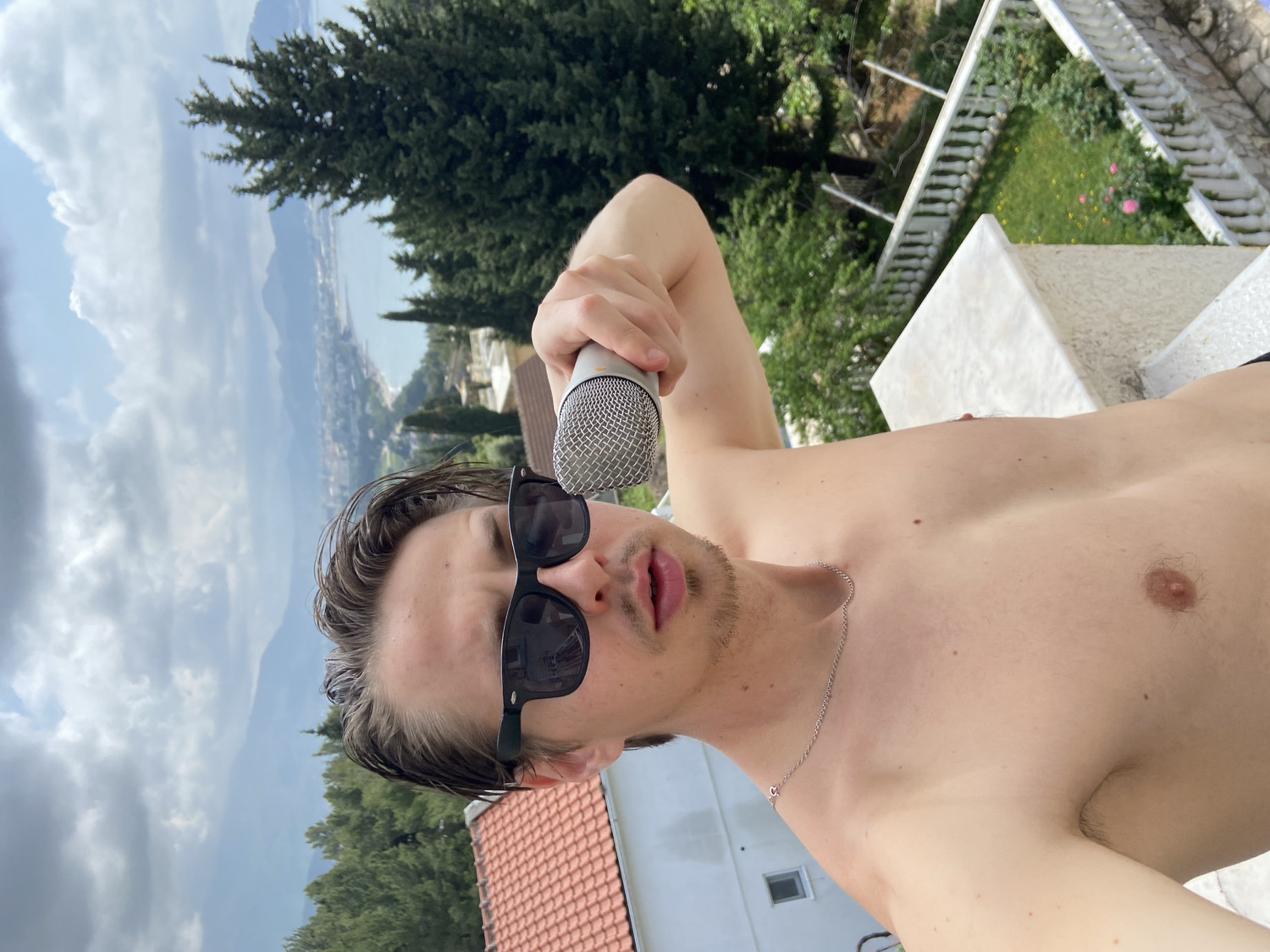 A young man is captured in a rotated, selfie-style image, appearing shirtless and wearing black sunglasses. He holds a silver microphone close to his slightly open mouth with his right hand, suggesting he is singing or speaking into it. A thin silver chain is visible around his neck. His hair is dark and styled back.
The background presents a scenic view under a bright, partly cloudy sky. Prominent dark green conifer trees frame the upper and middle ground. Beyond them, a coastal town stretches along a body of water, likely a bay or the Adriatic Sea, with distant mountains completing the horizon. Below, part of a white building with a red tiled roof is visible, along with a white railing or balustrade that hints at a balcony or terrace setting. The weather is sunny with scattered white clouds. No text is visible in the image. The landscape is consistent with the coastal region of Šušanj, Montenegro.
