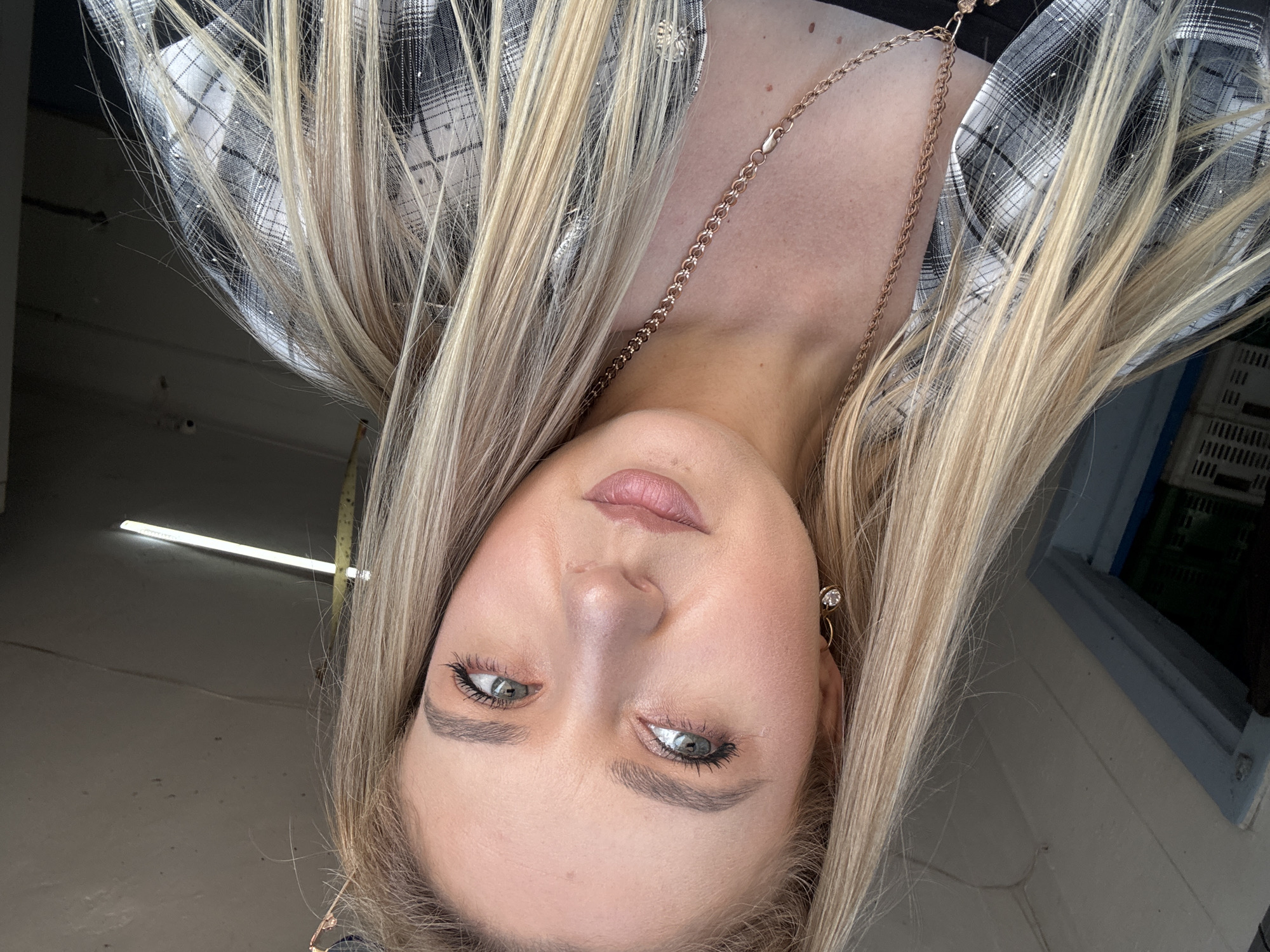 A blonde woman with blue-green eyes is pictured from the chest up, looking slightly to her left. She has on a black and white plaid shirt over a black top, and is wearing a gold chain necklace with a pendant. The background appears to be an indoor or covered area with concrete walls and a fluorescent light fixture. The lighting is bright, suggesting daytime.
