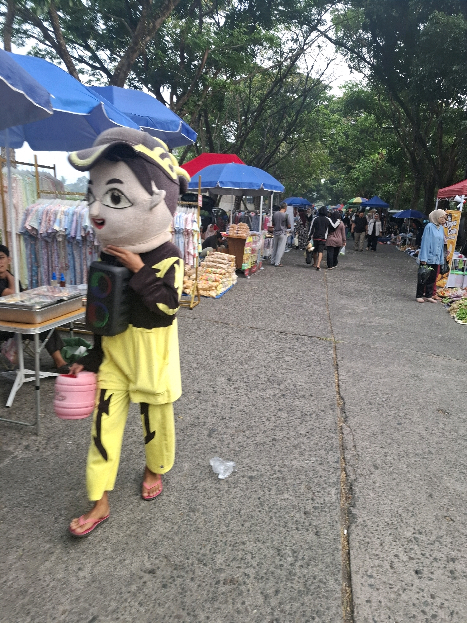 A person wearing a mascot costume is walking on a paved road in an outdoor market. The costume features a large, cartoonish head with wide eyes and an open mouth, a dark brown jacket with yellow accents, and bright yellow pants. The person is carrying a black portable speaker in their right hand and a pink bucket in their left. They are wearing pink flip-flops. In the background, there are market stalls with various goods displayed under blue and red umbrellas. Several other people are browsing at the market. The environment appears to be a street lined with trees, suggesting an outdoor setting in Karangtengah, Indonesia.