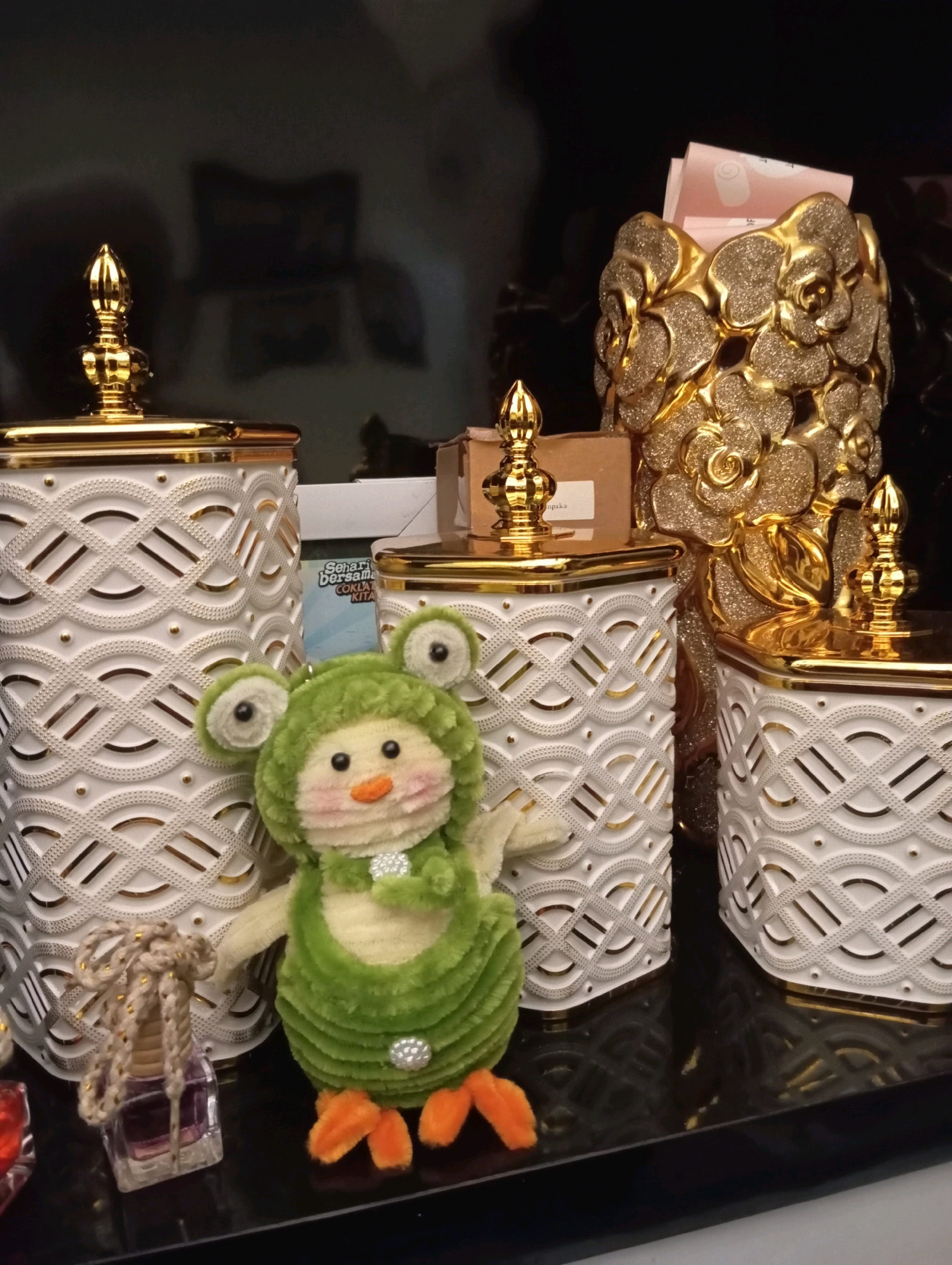 This image depicts a collection of decorative objects, including ornate gold-accented white jars, a gold rose-shaped vase, and a plush green frog toy, displayed on a reflective black surface. A small purple bottle with a rope detail is also visible. The setting does not provide any information about its location in Garut, Indonesia, nor does it show any people or actions.