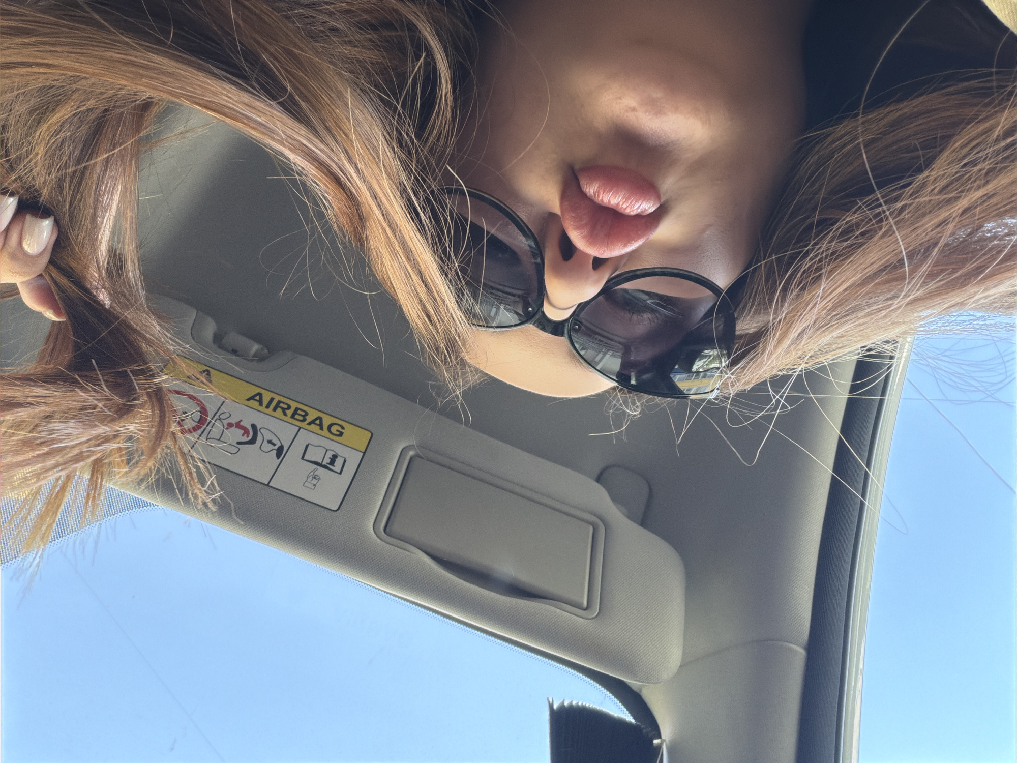 The image shows a selfie taken inside a car. The individual is positioned with their face tilted downward towards the camera. They are wearing sunglasses and sticking their tongue out. A portion of the car's interior is visible, including the sun visor with an "Airbag" warning label. Outside the car window, there's a view of a clear blue sky. There is no evidence to support the claim that the location is in the city of Chaikovsky, Russia.