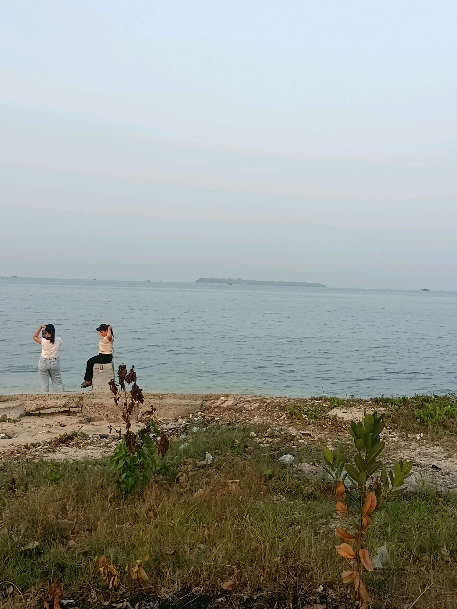 The image depicts a coastal scene with two women by the water. One woman, standing on the left, is wearing a white t-shirt and light blue jeans, holding her hair up. The other woman, seated on a concrete structure on the right, is wearing a striped t-shirt and dark pants, also with her hands near her hair. The water is calm, with a distant island or landmass visible on the horizon. The foreground shows a rough, somewhat unkempt shoreline with dry vegetation, scattered debris, and concrete structures. The sky is overcast with a pale, hazy appearance. The scene appears to be in a coastal area, possibly a less developed or natural part of Cikal, Indonesia.