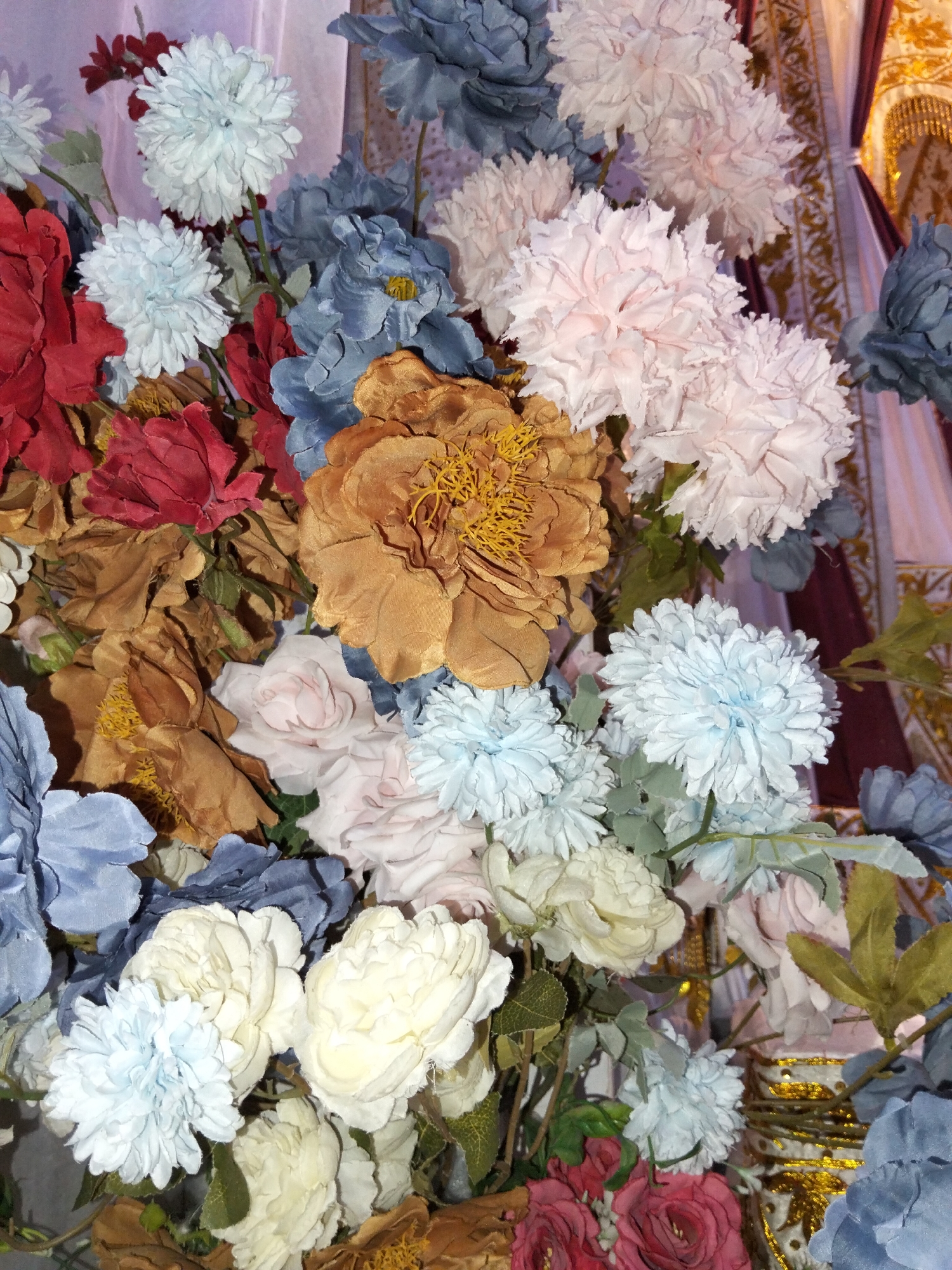 This is a close-up, eye-level, indoor photograph of a dense arrangement of artificial flowers. The flowers are rendered in various shades of light blue, dusty rose, muted gold, white, and deep red, with some foliage interspersed. The textures are varied, with some flowers having delicate, ruffled petals, while others have more structured, layered forms. The lighting is even and diffused, suggesting an indoor setting, possibly a studio or event space. The background is out of focus, showing hints of draped fabric in muted tones of pink and gold, and a decorative element that suggests an ornate setting. There are no people or animals visible in the image, and no specific geographic markers are present to indicate a location within Cottrueng, Indonesia, or any other place.