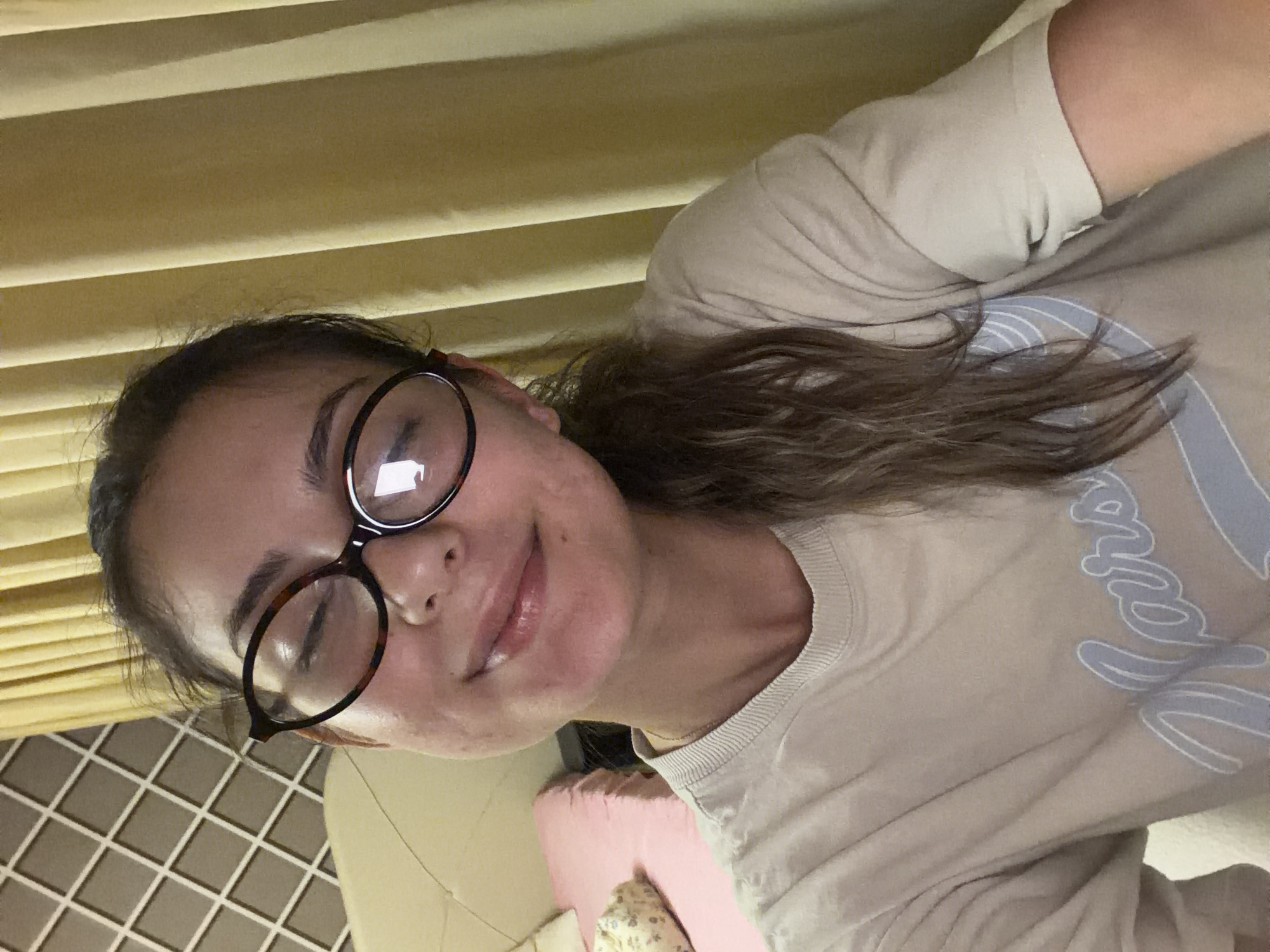A young woman with brown hair and glasses is taking a selfie. She is wearing a light-colored t-shirt with a blue design. The background shows a wall with a patterned design and yellow curtains. A pink pillow and part of a bed are visible to the left. The lighting is dim, suggesting an indoor setting, possibly a bedroom. There is no specific indicator of the city of Burgas, Bulgaria, in the image.