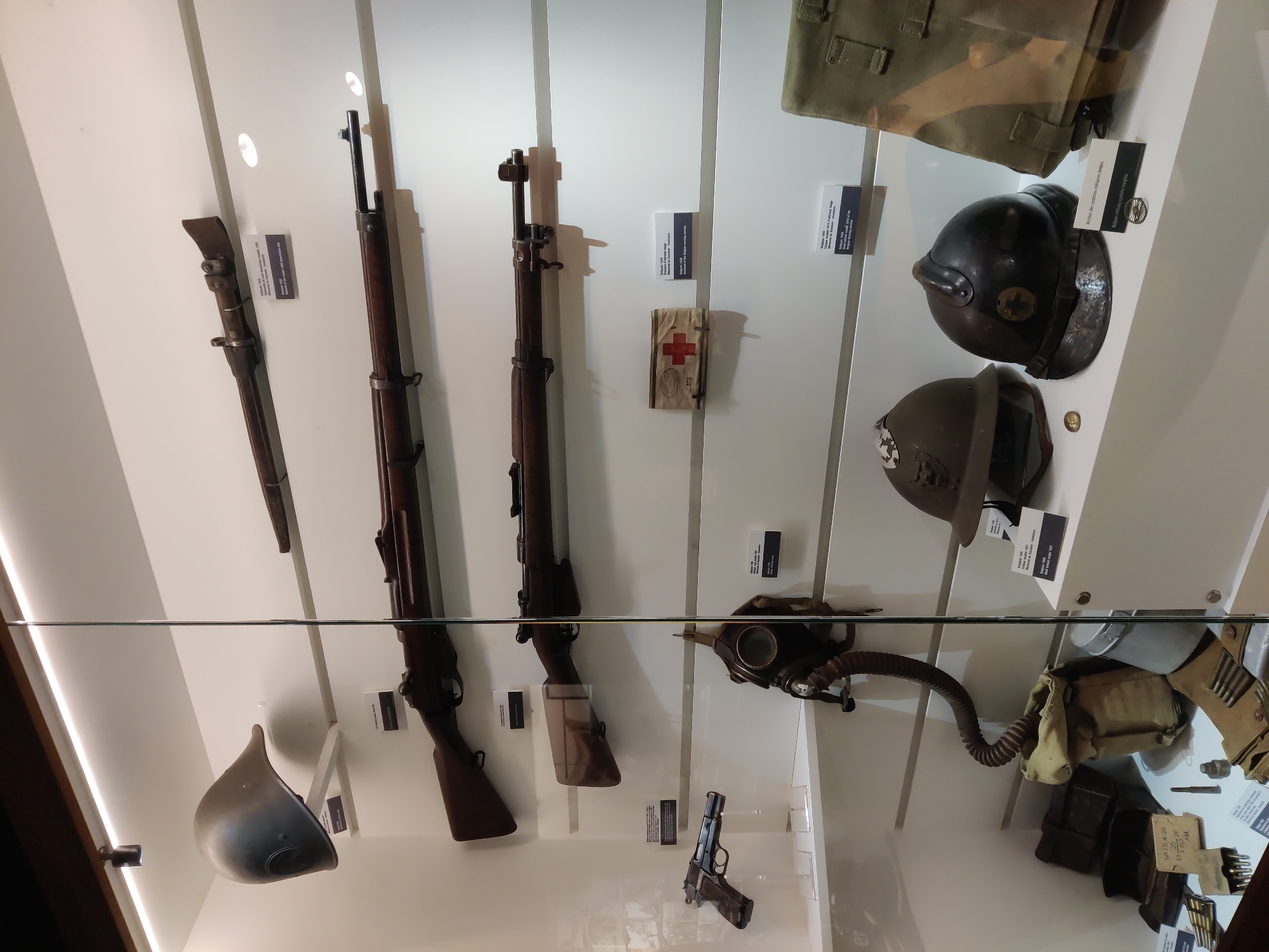 The image is a close-up, eye-level, horizontal shot of a museum display case containing military artifacts, likely in Dunkirk, France, given the items' association with that region. The display features several rifles mounted horizontally on white shelves, with accompanying informational placards. A helmet is displayed on the top shelf to the left. Below the rifles, a pistol is mounted on a clear stand. A gas mask with a coiled hose hangs on the right side of the lower shelves. Further down, a collection of smaller items, including more helmets, pouches, ammunition, and a metal container, are arranged. The lighting is bright and evenly distributed from above, illuminating the artifacts and casting subtle shadows. The overall environment appears to be a clean, well-organized museum exhibit, enclosed by glass.