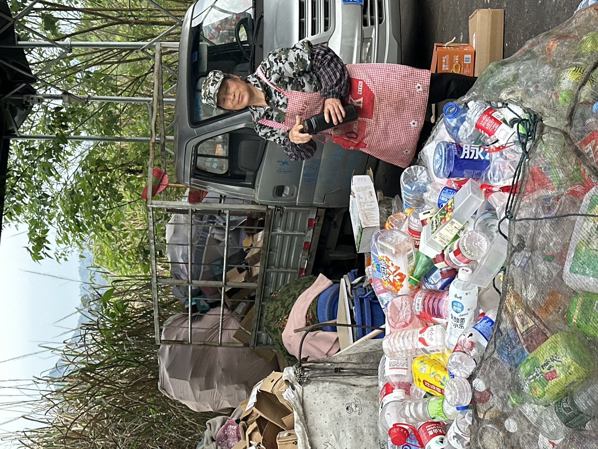 A woman in Jiangjin, China stands next to a silver truck laden with recycled goods. She is wearing a camouflage jacket, a cap, and a red-and-white checkered apron. She is holding a black thermos in her hands. Next to her is a large pile of plastic bottles contained in a mesh bag. Behind the truck is a metal frame filled with various items, including cardboard boxes and cloth. Tall grasses are visible in the foreground.