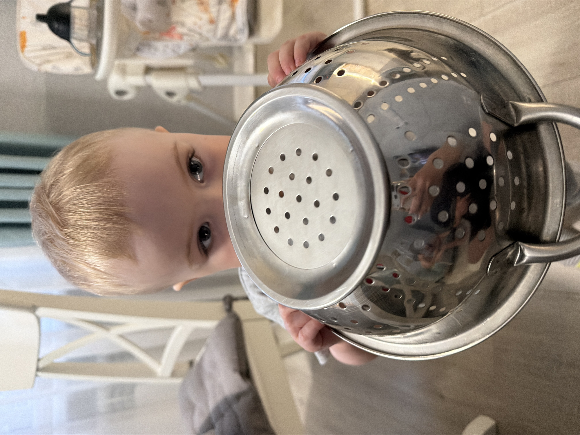 A child with blond hair is holding a stainless steel colander in front of their face, obscuring most of their features except for their eyes and a portion of their forehead. The colander is positioned in the foreground, with the perforated base facing the viewer. The background includes a white high chair with baby supplies. The location is likely indoors on a light brown floor. There is no indication of the city of Votkinsk, Russia.