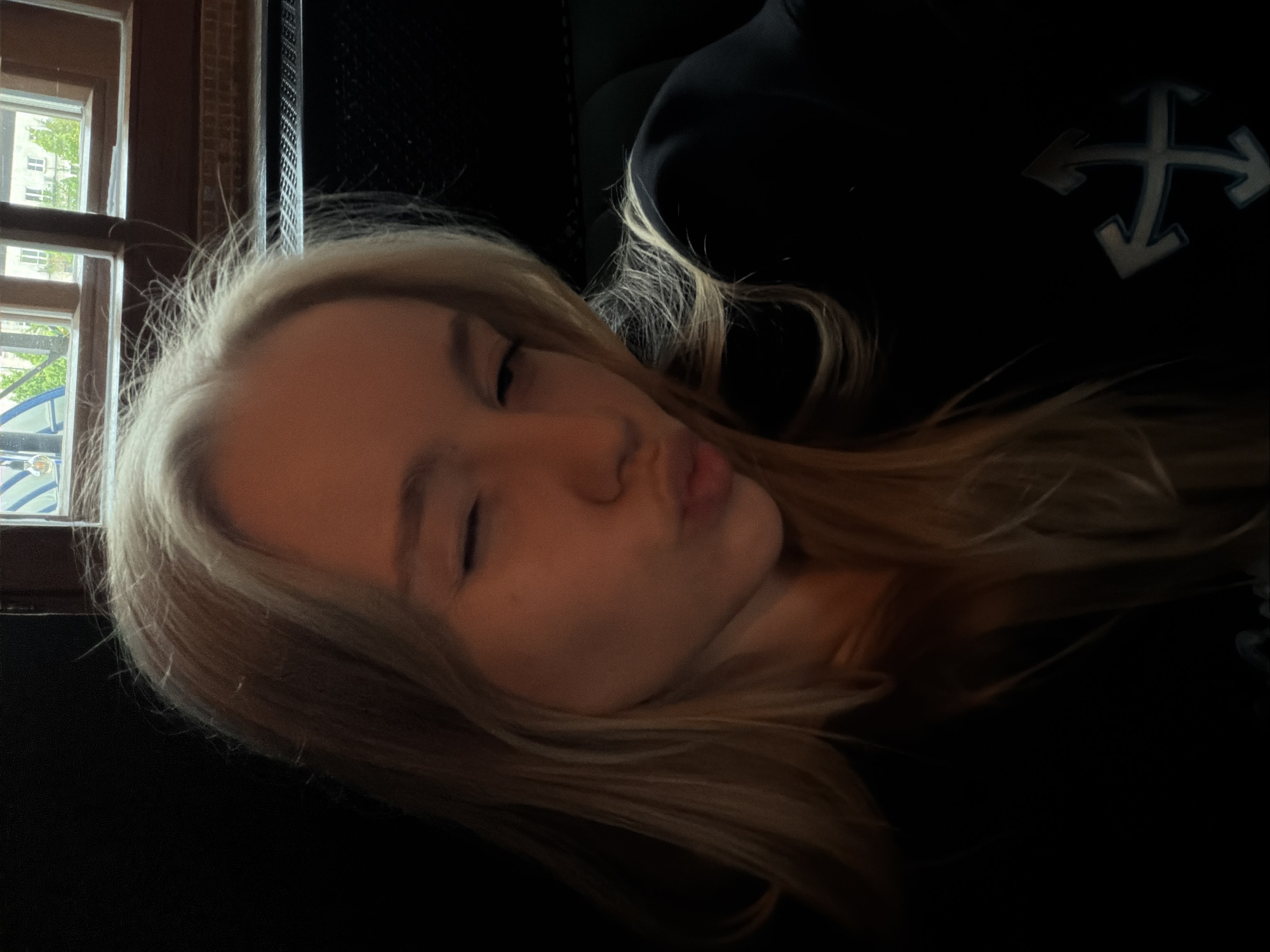 A young woman with blonde hair is reclining indoors. She is wearing a black sweatshirt with a white four-way arrow symbol on the upper right side of the frame. Her eyes are partially closed and she is puckering her lips. A window with a partially obscured view of buildings and trees can be seen to the left of her head. The location is Unknown, Unknown.