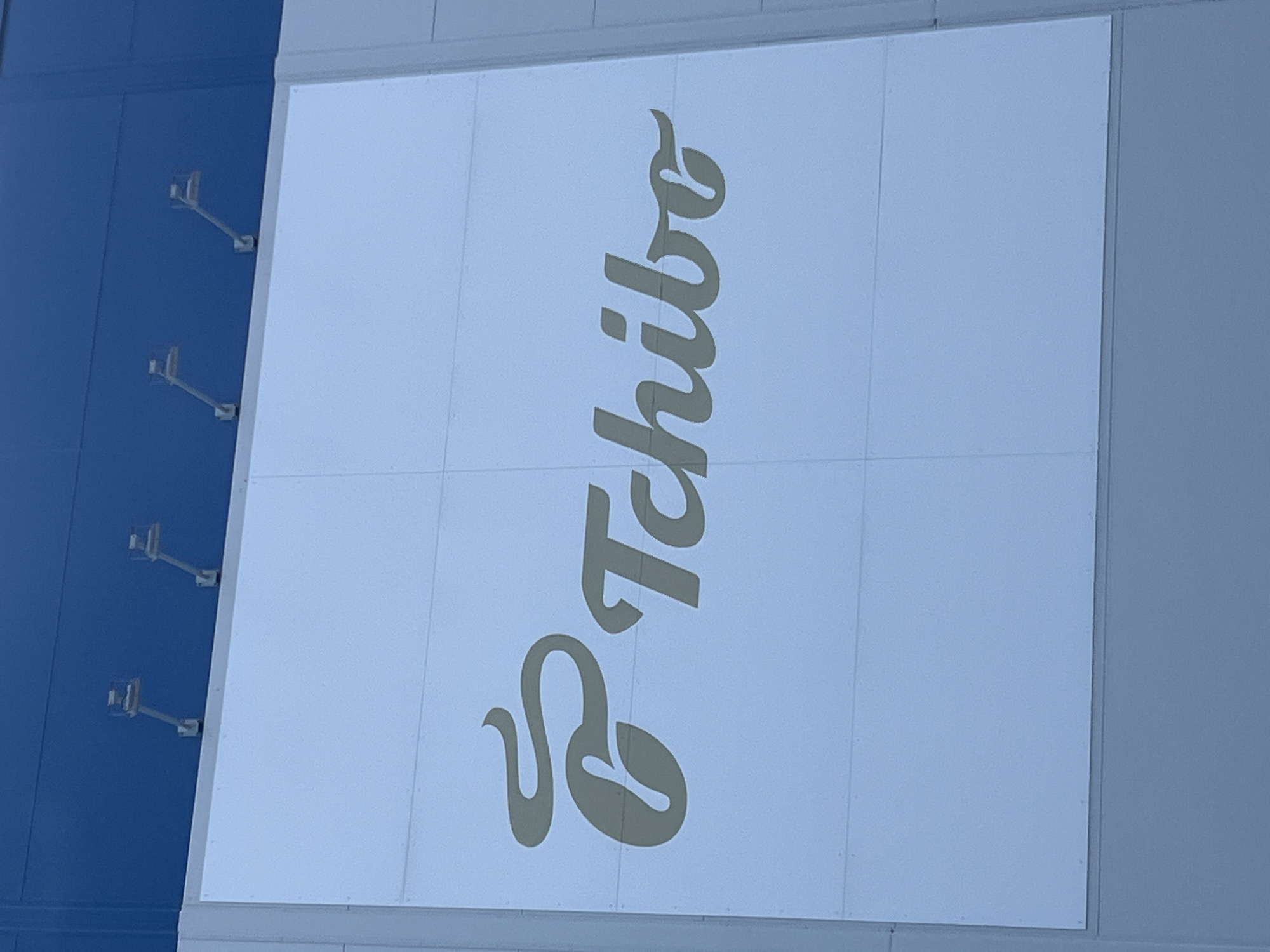 The image displays a building facade with a large sign featuring a logo and text. The sign is light-colored and appears to be mounted on the building's exterior. Above the sign, there are several metal fixtures attached to the dark blue upper portion of the building. The text on the sign reads "Tchibo." Based on the context provided, this image is located in the city of Cheb, Czechia. The scene is static, with no visible people or ongoing actions. The environment appears to be outdoors during the day, given the lighting.