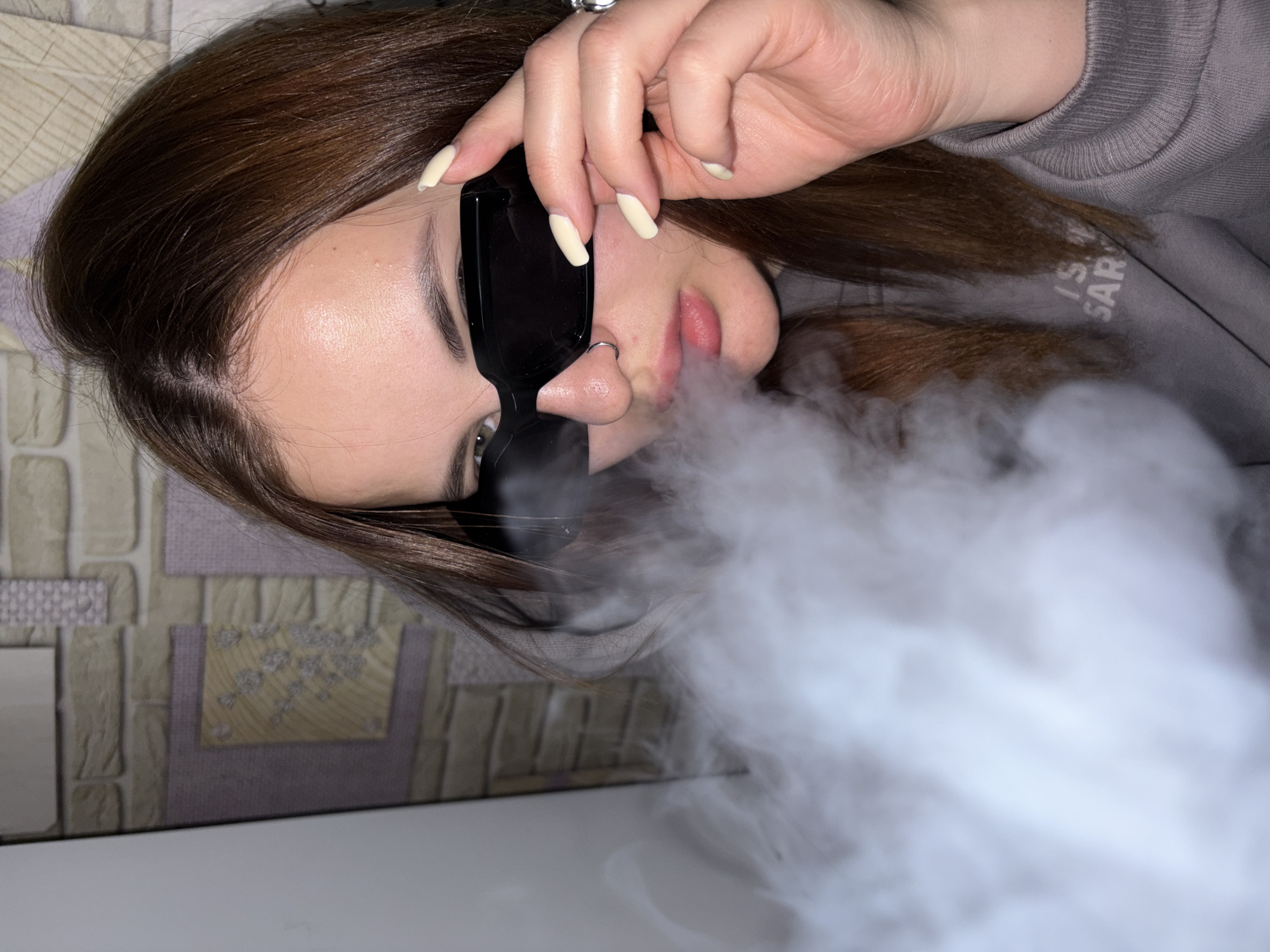 A young woman is captured in a close-up, rotated portrait, exhaling a dense cloud of white vapor or smoke. She is positioned diagonally in the frame, looking towards the viewer with her head tilted to the side. Her face is partially obscured by large, dark sunglasses which she holds onto with her right hand, revealing long, cream-colored, manicured nails. A silver ring is visible on her finger. She has long, dark brown hair parted on the side. Her skin appears fair, and she has a subtle nose ring on her left nostril. Her lips are slightly parted and pink.
She is wearing a gray sweatshirt, and faint text, possibly "IS.W SAR" or similar, is visible on the fabric near her shoulder. The background consists of a patterned wall, appearing to be wallpaper with various rectangular sections, some depicting a brick-like texture and others with decorative motifs, suggesting an indoor setting. Below her, a plain white surface is visible, possibly a table or ledge. The overall mood is casual and stylish, with the smoke creating a dramatic effect. The location context for this image is Port-Katon, Russia.