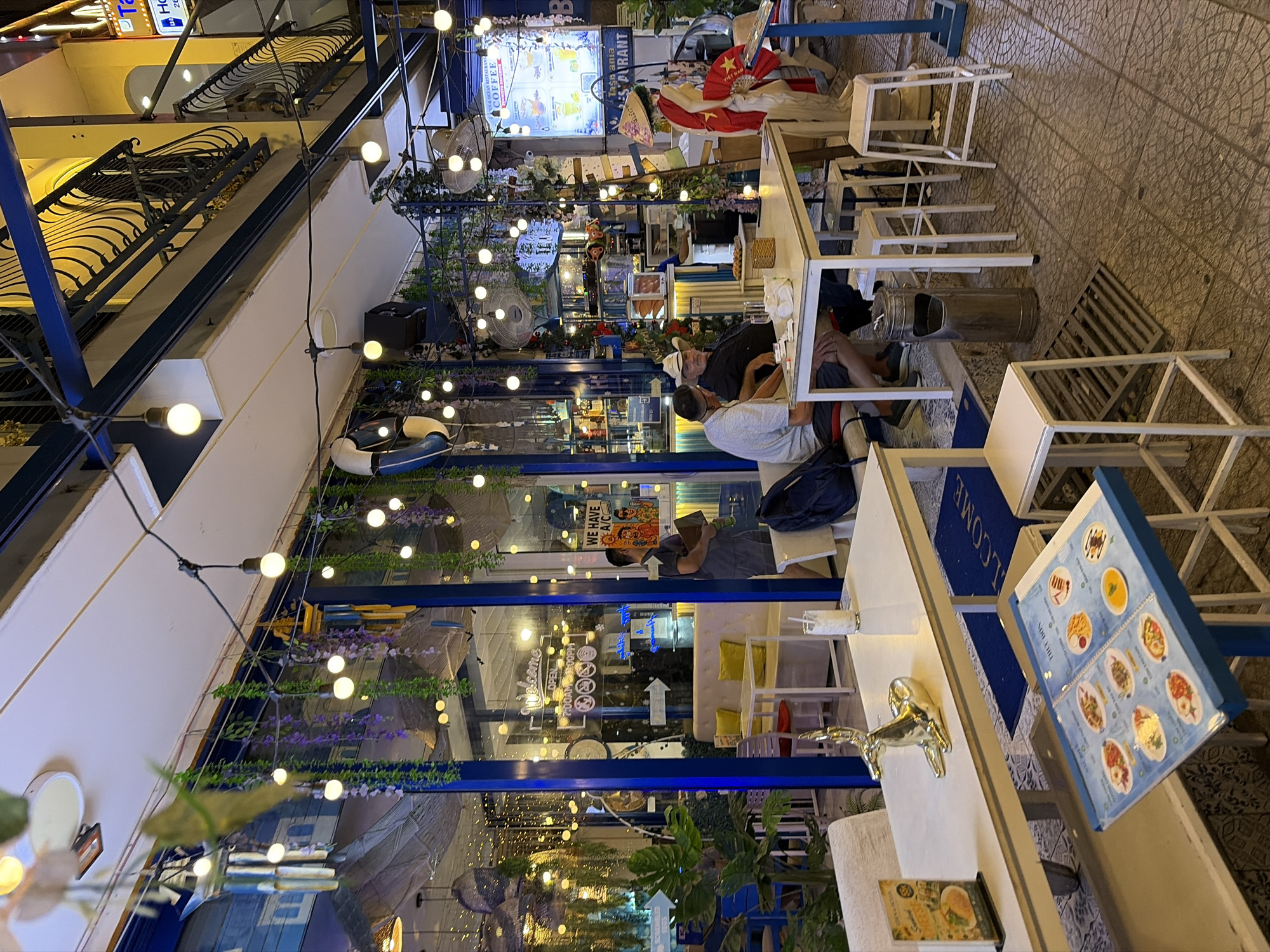 The image depicts the outdoor seating area of a restaurant named "BLUE OCEAN RESTAURANT" located in the city of Đà Nẵng, Vietnam. The restaurant has a vibrant, blue-themed facade with large glass windows. The outdoor space is decorated with numerous strings of fairy lights and hanging plants, creating a festive atmosphere.
Several patrons are visible. Two men, one wearing a white cowboy hat and the other in a grey t-shirt, are seated at a white table, engaged in conversation. To the right, a person dressed in a traditional Vietnamese conical hat and a red garment stands near a decorative mannequin. Inside the restaurant, through the glass windows, a few other individuals can be seen, and a seating area with yellow cushions is visible.
Objects of note include white tables and chairs, a metal ashtray, and a menu displayed on a stand in the foreground. The ground is paved with patterned tiles. The lighting suggests it is either dusk or early evening, as the artificial lights are prominent. The overall scene conveys a relaxed and inviting dining environment.