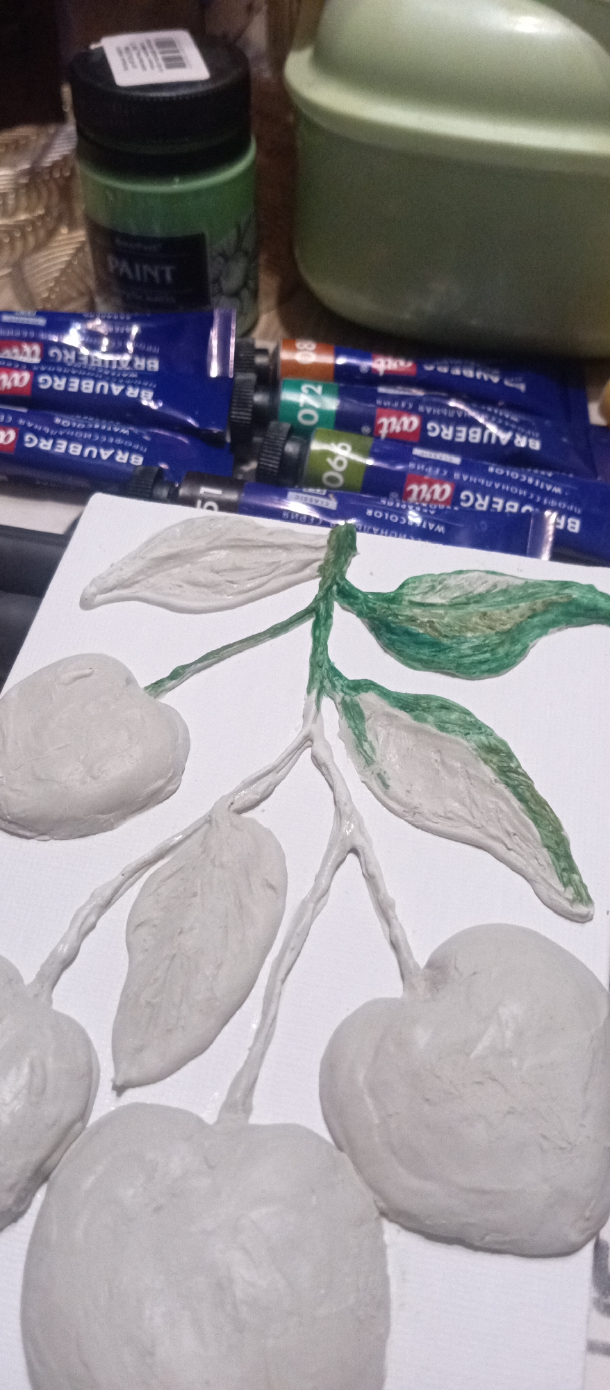 The image shows an unfinished artwork with clay apples and leaves on a white background. Some parts of the leaves and stems have been painted green. Tubes of Brauberg watercolor paints in shades of green, blue, and brown are scattered near the artwork. A jar of green paint and a green container are placed in the background.