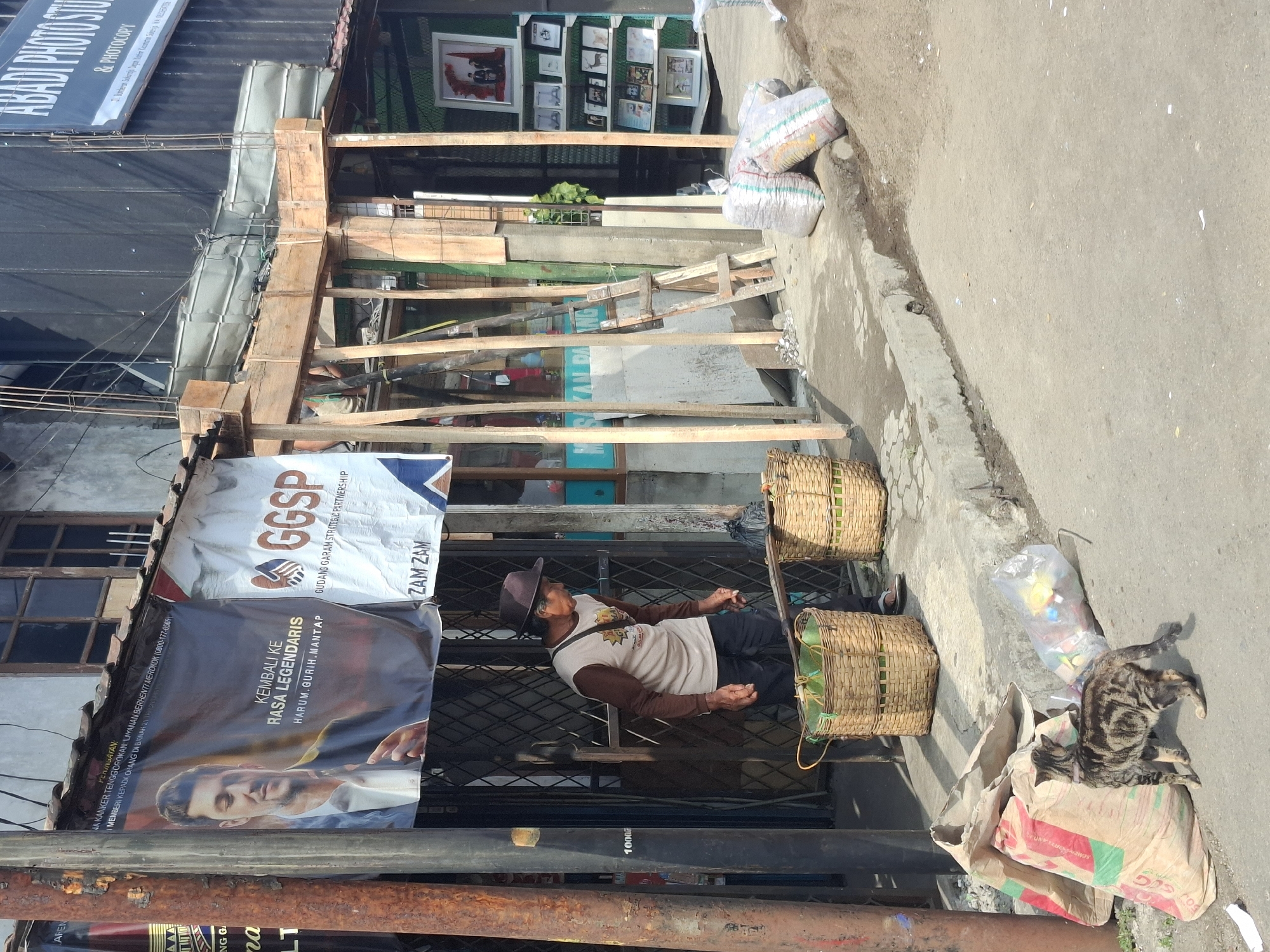 This is a medium shot image of a street scene in Sukaraja, Indonesia, on a bright, sunny day. In the foreground, a tabby cat is seen from behind, looking towards a large pile of sacks and a clear plastic bag filled with colorful objects, situated on a paved surface. Further back, a man wearing a hat and a light-colored long-sleeved shirt with dark pants is standing and looking down. He is positioned between two large woven baskets, one of which appears to be filled with green leafy produce.
Behind the man and baskets, a building is visible, featuring a dark-colored facade with metal grating and signs displayed. A prominent banner with an image of a man and text that reads "KEMBALI KE RASA LEGENDARIS HARUM.GURIH.MANTAP" is hanging from the building's overhang. Another banner, white with red and blue graphics, reads "GGSP GUDANG GARAM STRATEGIC PARTNERSHIP".
The architecture suggests a commercial or workshop area, with wooden framing and metal supports indicating ongoing construction or renovations. Various items are scattered on the ground, including smaller sacks, debris, and a metal pole. The ground surface varies from paved areas to dirt and gravel. A ladder leans against the wooden structure. In the background, framed pictures are displayed on shelves. The overall impression is a lively street scene in a commercial district.