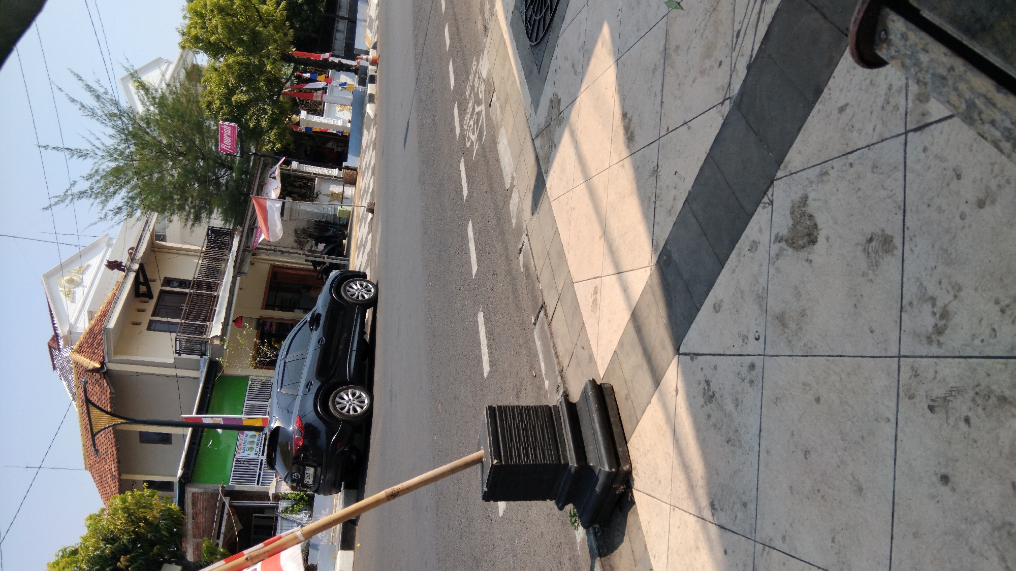 The image depicts a street scene in Batang, Indonesia, on a clear day. In the foreground, a concrete sidewalk is visible, paved with square tiles, some of which show marks and stains. A black, decorative post with a wooden pole inserted into it stands on the edge of the sidewalk, near the street.
The street itself is asphalt, with white dashed lines indicating lanes. A black SUV is parked on the side of the road. Behind the car, a two-story building with a red-tiled roof is visible. The building has balconies and windows, and an awning or canopy extends over part of the lower level. A sign with the word "Flourish" in pink is attached to the building.
To the right of the building, there are trees, and a series of flags, some red and white, are displayed. The general environment suggests a commercial or residential area within the city. The time of day appears to be morning or afternoon, given the presence of shadows cast by the sun.
