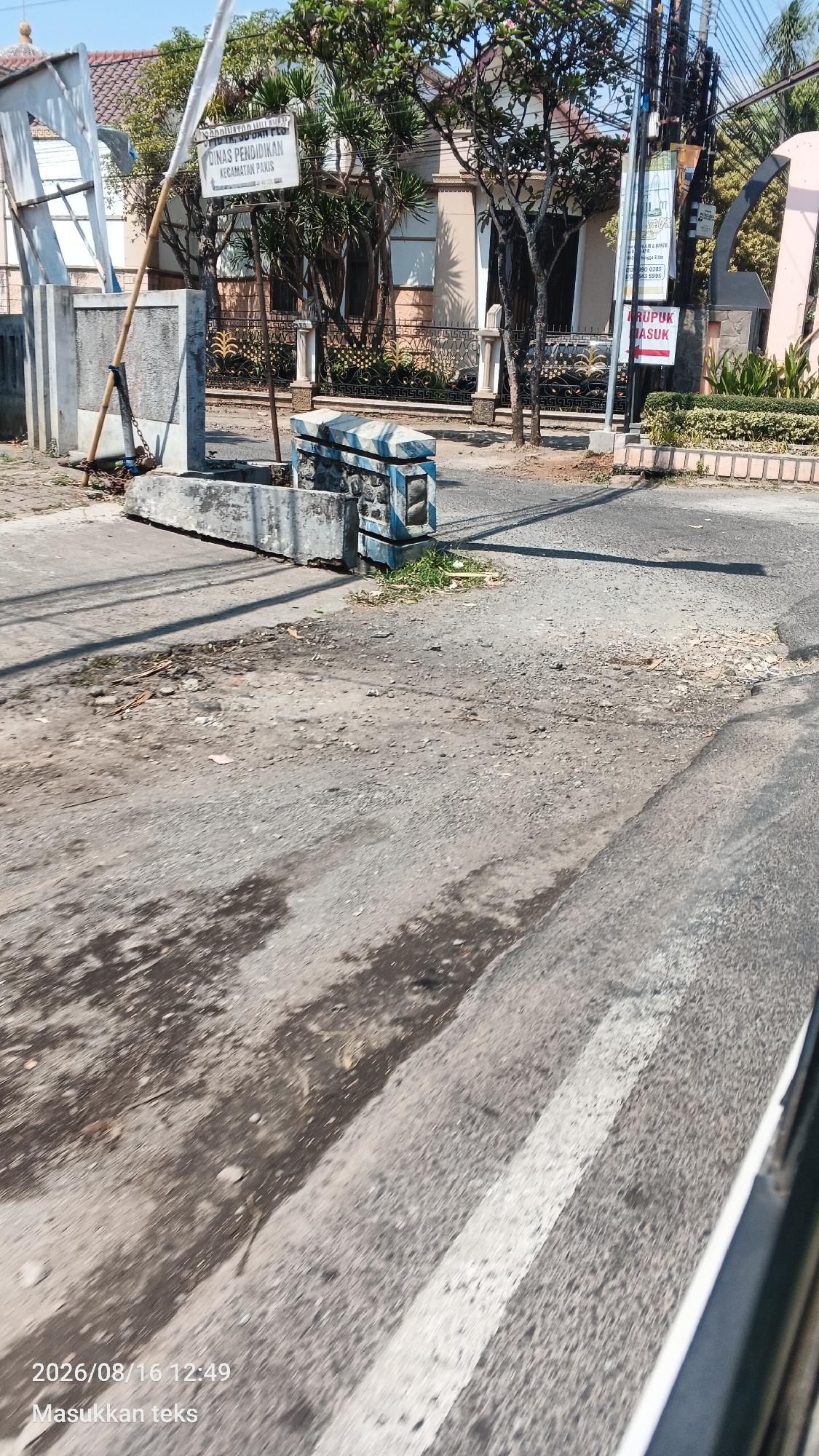The image depicts a street scene in front of a building with a sign reading "DINAS PENDIDIKAN KECAMATAN PAKIS", indicating it is the District Education Office in Pakis, Indonesia. The foreground shows a cracked and weathered asphalt road with some debris scattered on it. A white line is painted on the road, suggesting a crosswalk or lane marking.
To the left, there is a concrete barrier with a metal pole attached, possibly part of a gate or barrier system. A white flag or banner is tied to the pole. Behind the barrier, a building with a tiled roof and light-colored walls is visible, partially obscured by trees and foliage. A black metal fence with decorative elements runs along the property line.
On the right side of the image, there are several utility poles with wires. Another sign is attached to one of the poles, with some text that is partially legible. Further to the right, there is a large pink arch-shaped structure and manicured green bushes.
In the middle ground, there is a low concrete structure decorated with blue and white striped bricks, with grass growing around it. The overall environment appears to be a public space or entrance to an administrative building. The image is dated 2026/08/16 12:49.