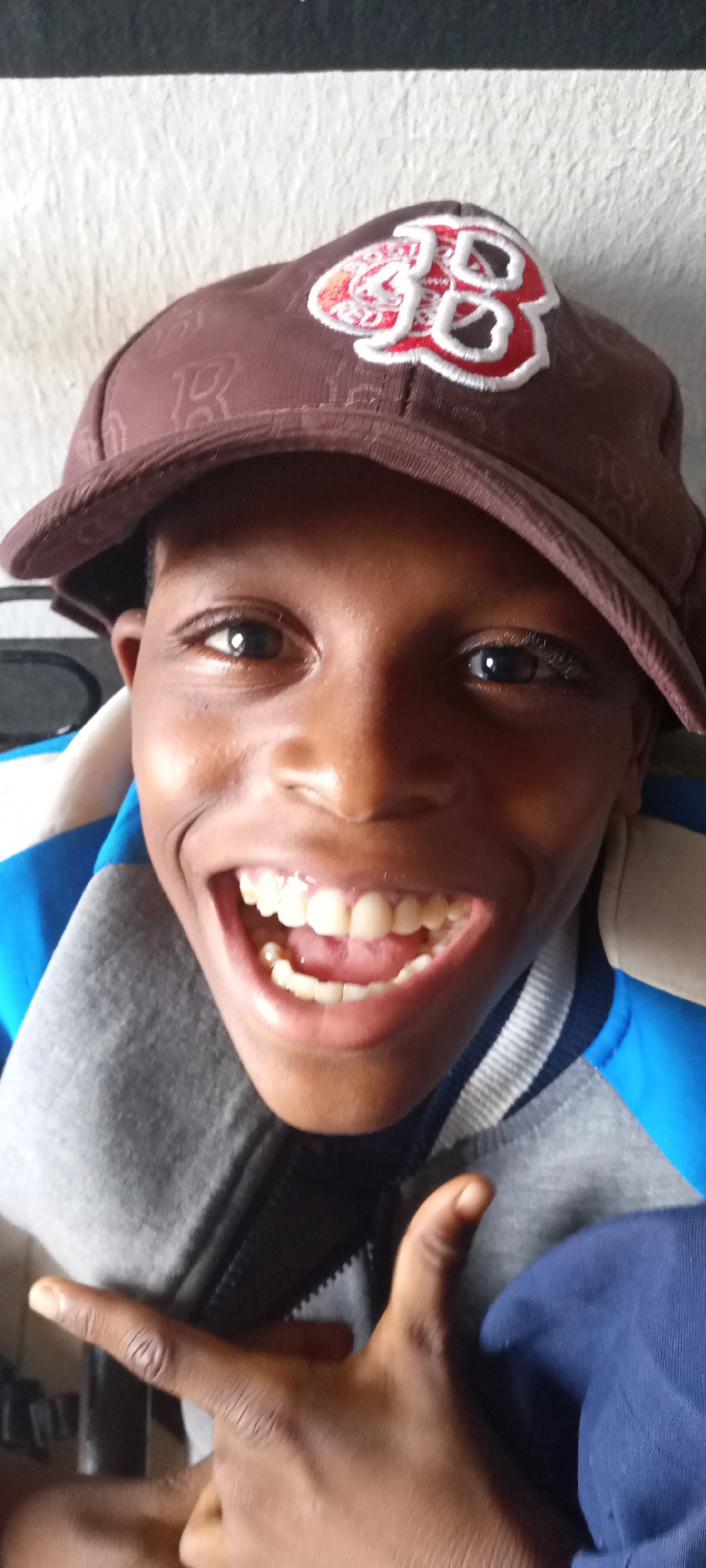 A young, dark-skinned person is seen from the chest up, smiling broadly with teeth visible. The individual is wearing a brown cap with a stylized "B" logo embroidered in red and white, a grey and blue jacket. One hand with darkened fingernails is visible, partially in front of the person’s chest. The background is a plain white surface. There are no indicators to determine if the picture was taken in Jos, Nigeria.