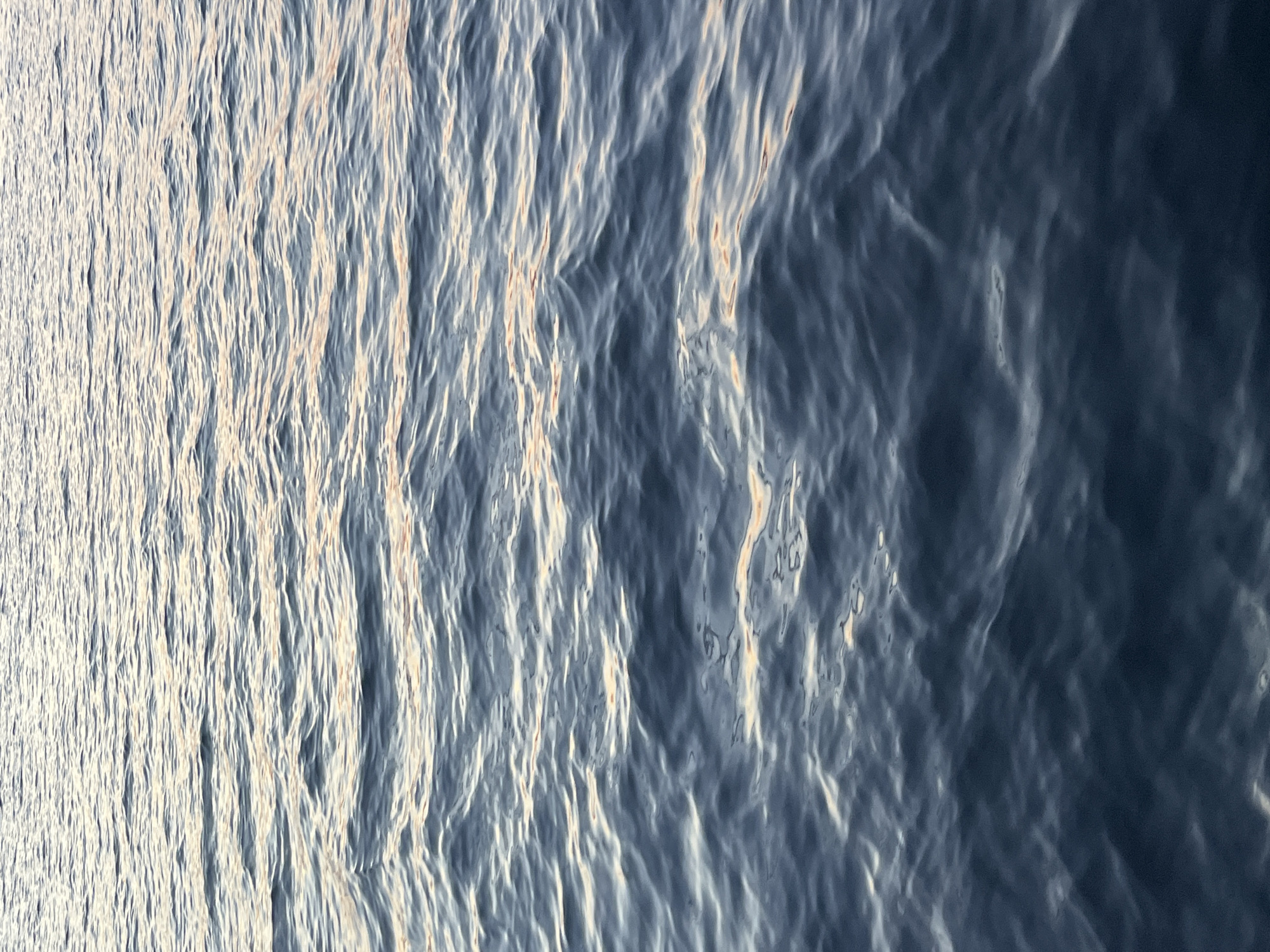 The image shows the surface of a body of water, possibly the Kama River. The water is rippled and varies in color from light grey to dark blue, suggesting varying depths or reflections. The image is taken from an elevated position, looking down at the water. There are no people, boats, or other objects visible on the surface. The lighting seems soft, indicating diffused sunlight. The consistency and movement of the water surface are consistent with the river. The location of Chaikovsky, Russia, is relevant as it is situated on the Kama River, which makes this a plausible setting.