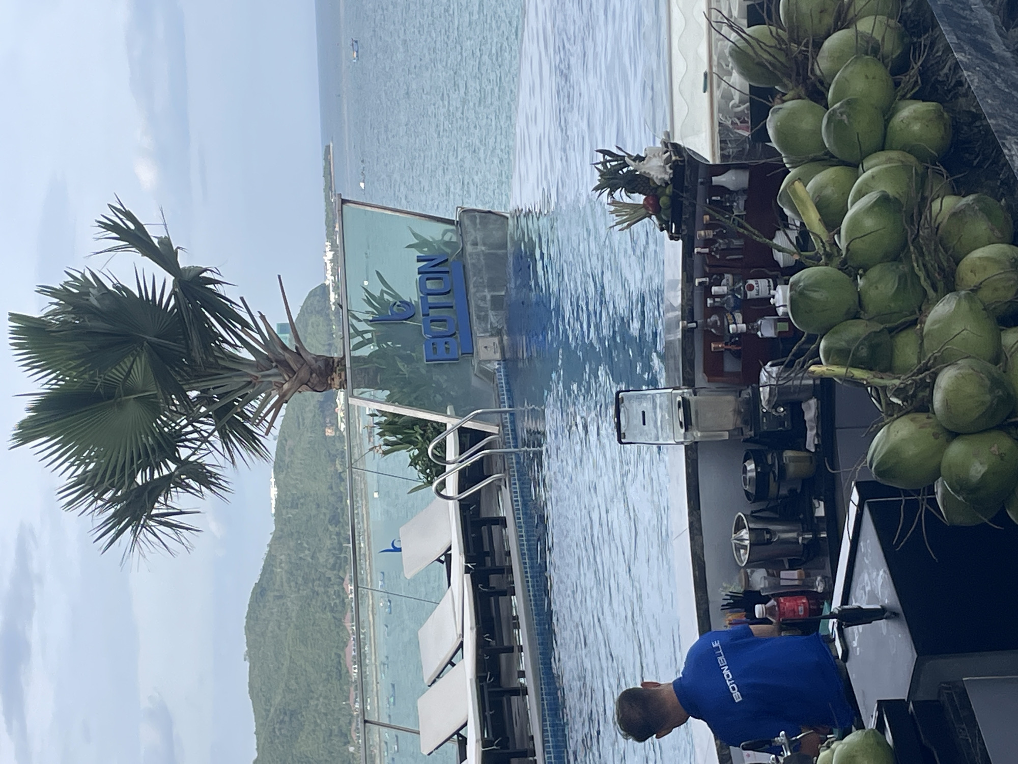 An exterior shot captures a resort with an infinity pool overlooking the ocean and a lush, green hillside. In the foreground, a bar area is stocked with coconuts, a blender, and various bottles, likely for drinks. A man in a blue shirt with "BOTON BLUE" printed on the back is tending to the bar. Beach chairs are arranged by the pool, and a palm tree stands tall against the cloudy sky. The "BOTON BLUE" logo is visible on the pool's edge. The scene appears to be located in Nha Trang, Vietnam.