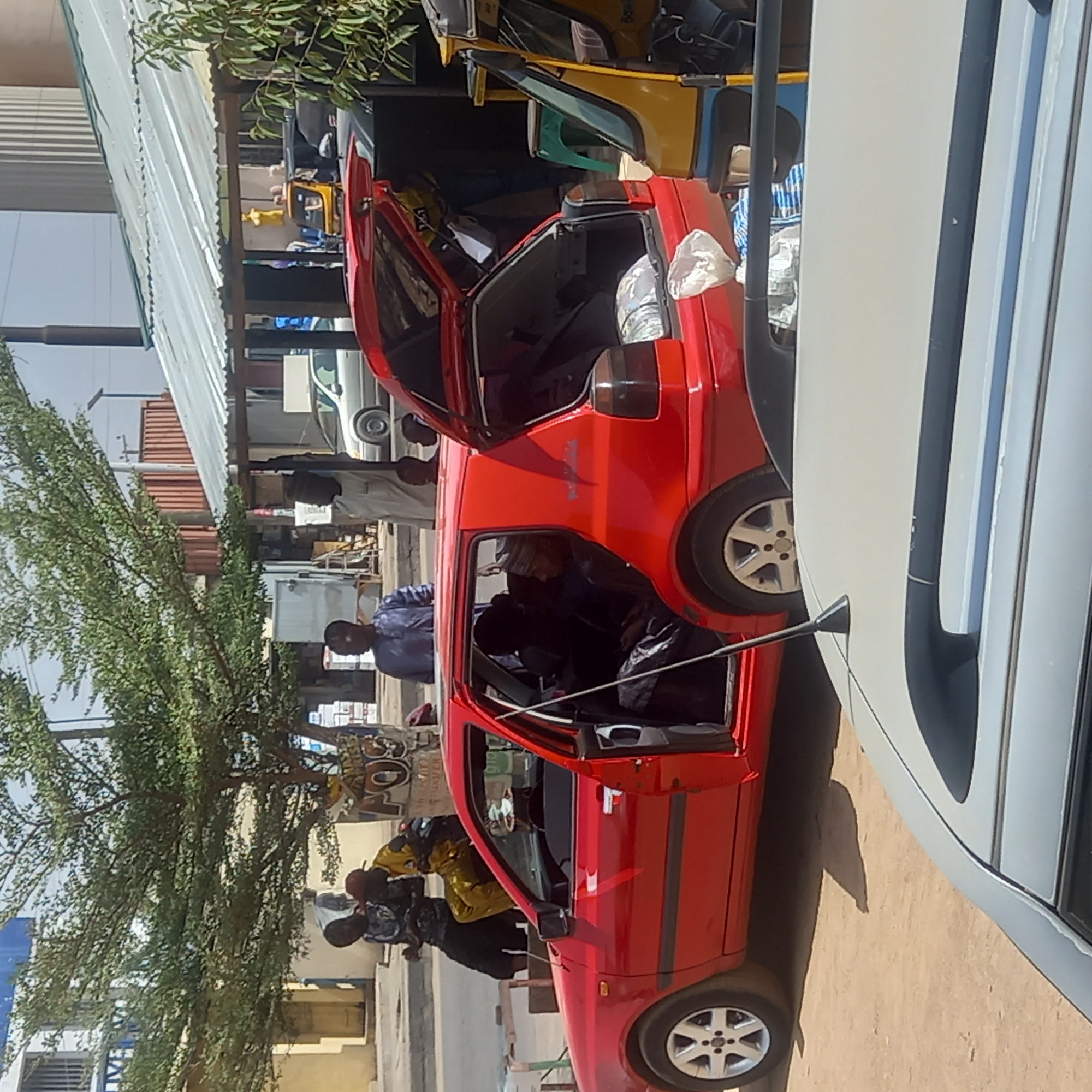 The image depicts a street scene in Dadi Riba, Nigeria. A red car has its doors open and parked on the side of the street. Individuals in casual clothing are standing near the car. Some other cars are visible in the background. There is a building and a tree.
