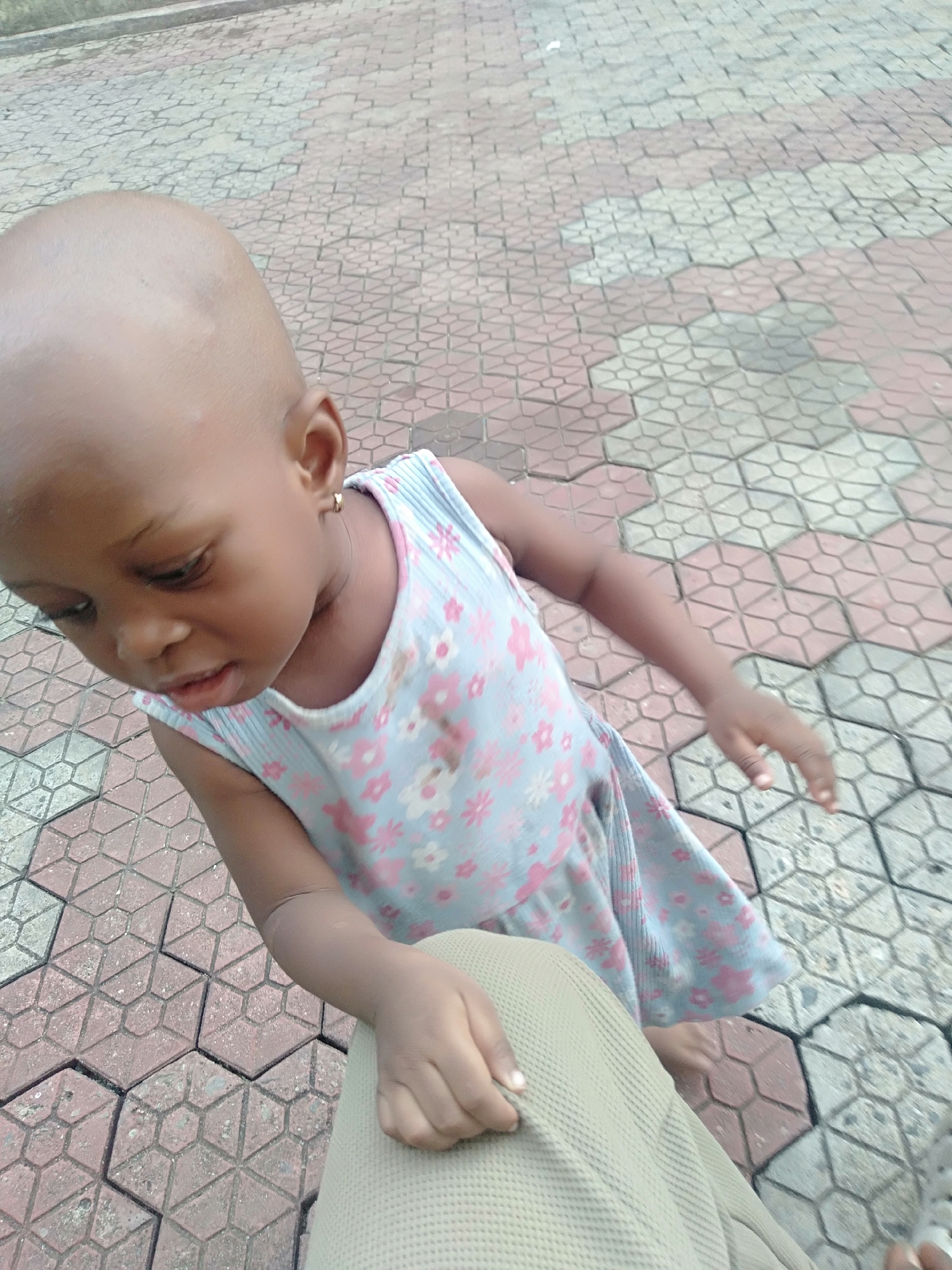 A young, bald child is seen outdoors on a patterned, tiled surface. The child wears a sleeveless, light blue dress with pink floral designs. The child is reaching towards a person wearing khaki-colored clothing. The location is likely Shangisha, Nigeria based on the prompt.