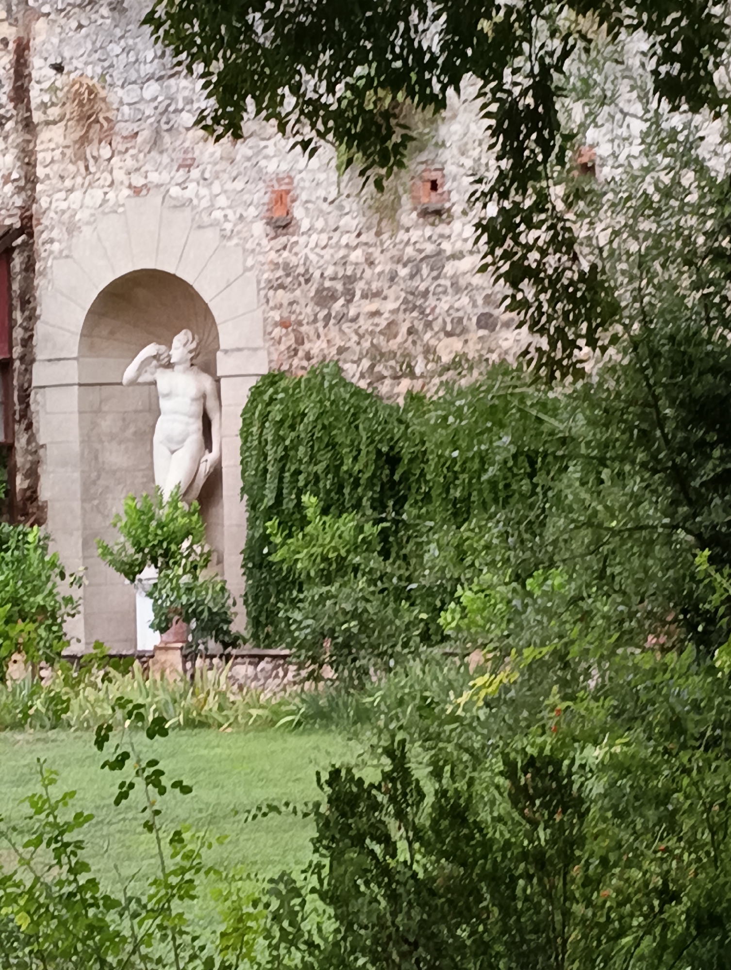 The image shows a stone sculpture of a nude male figure standing in an arched alcove against a rough-hewn stone wall. The sculpture is white and detailed, depicting the figure with one arm raised to his head and the other resting on his thigh. Lush green foliage, including a weeping tree and smaller potted plants, partially obscures the sculpture and the surrounding garden. A grassy lawn is visible in the foreground. This scene is located in Verona, Italy, likely in a garden setting.