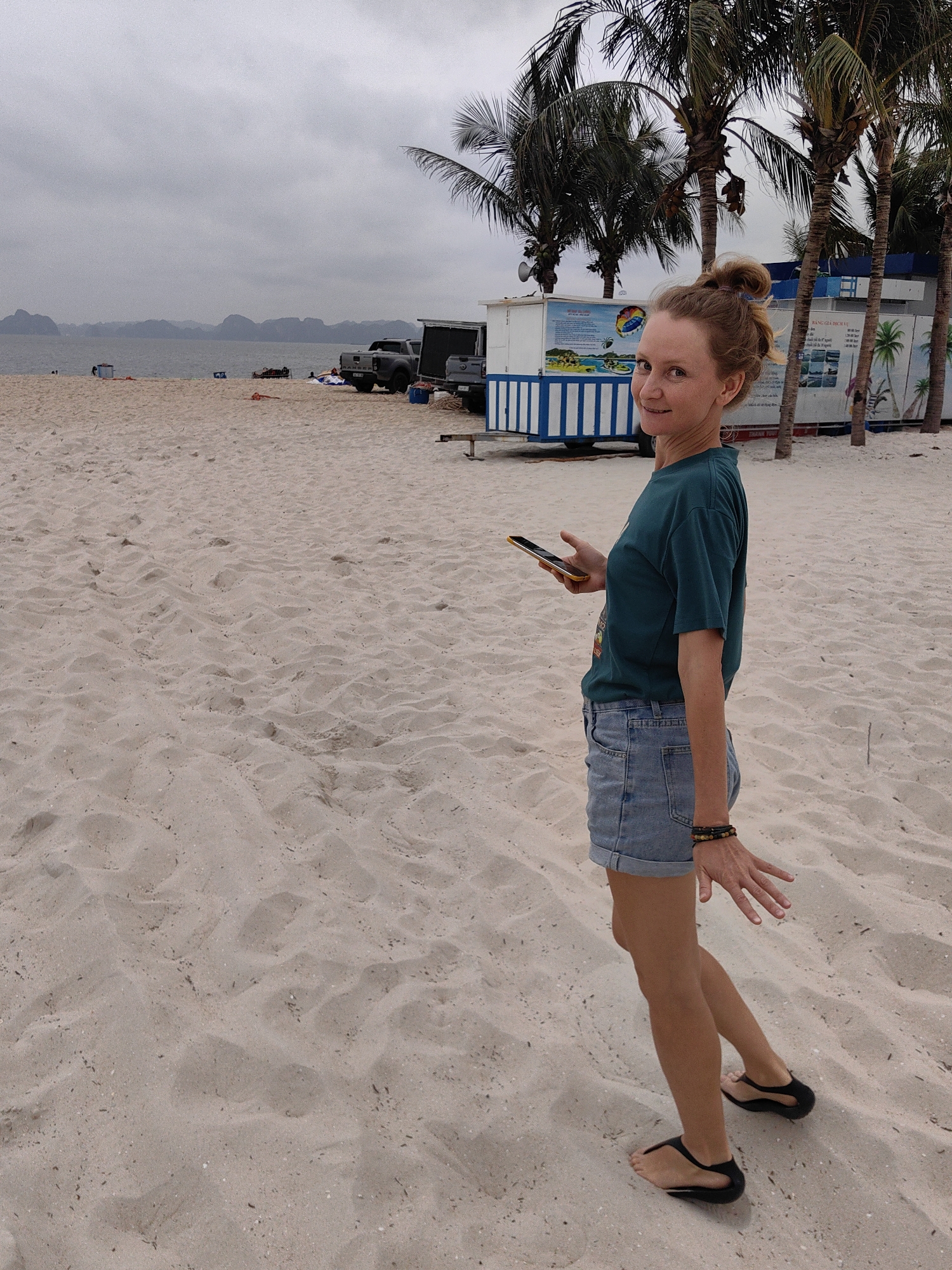 A woman with blonde-ish hair tied in a bun is captured mid-stride on a light-colored sandy beach in Bai Chay Ward, Vietnam, looking back over her right shoulder with a faint smile. She is dressed in a dark teal t-shirt, light blue denim shorts, and black flip-flops. In her left hand, she holds a yellow-cased smartphone, and a multi-beaded bracelet adorns her right wrist.
The beach is wide and expansive, with visible ripples and footprints in the sand. The background features a calm, grey sea under an overcast sky, with distinctive misty limestone karsts or mountains rising dramatically from the water in the distance.
Behind the woman, further along the beach, are several man-made structures and vehicles. A prominent white and blue striped shack, likely a service or information stand, displays signs with graphics and Vietnamese text, including "BANG GIA DICH VU" (Price List Service) and "BANG QUY DINH" (Regulations). Adjacent to it stands another, darker building. Two pickup trucks, one dark grey with "RANGER" visible on its tailgate, are parked on the sand near these structures. Tall palm trees line the area behind the buildings. The overall scene suggests a casual, cloudy day at a coastal tourist destination.
