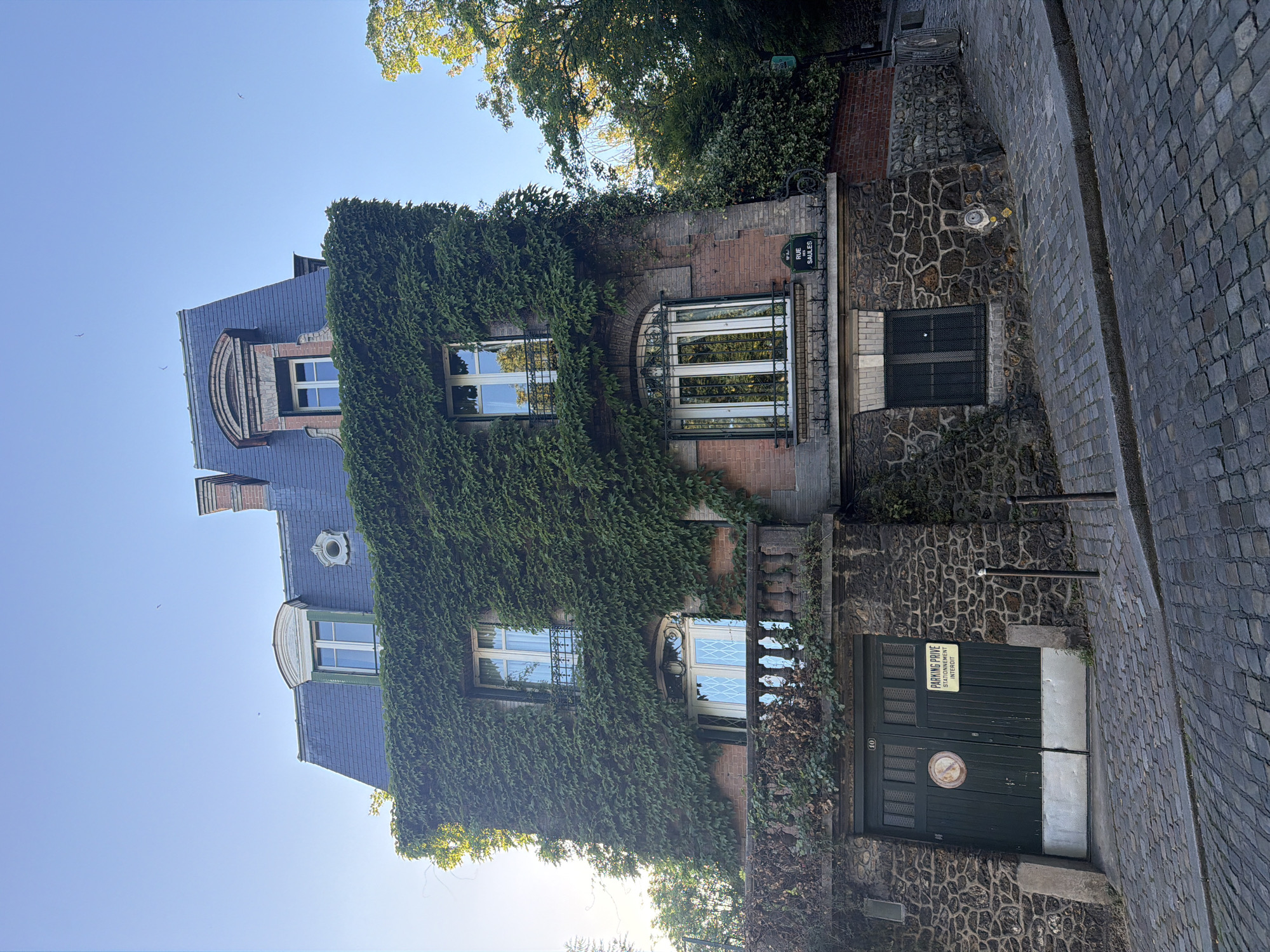The image depicts a multi-story building in Paris, France, heavily covered in ivy. The building has multiple windows and a dark-colored roof. A cobblestone street runs adjacent to the building. A sign indicating "Rue Sailles" is visible, confirming the location. A lower level of the building appears to have a gated entrance. The sky is light blue and some treetops are visible at the top of the image.