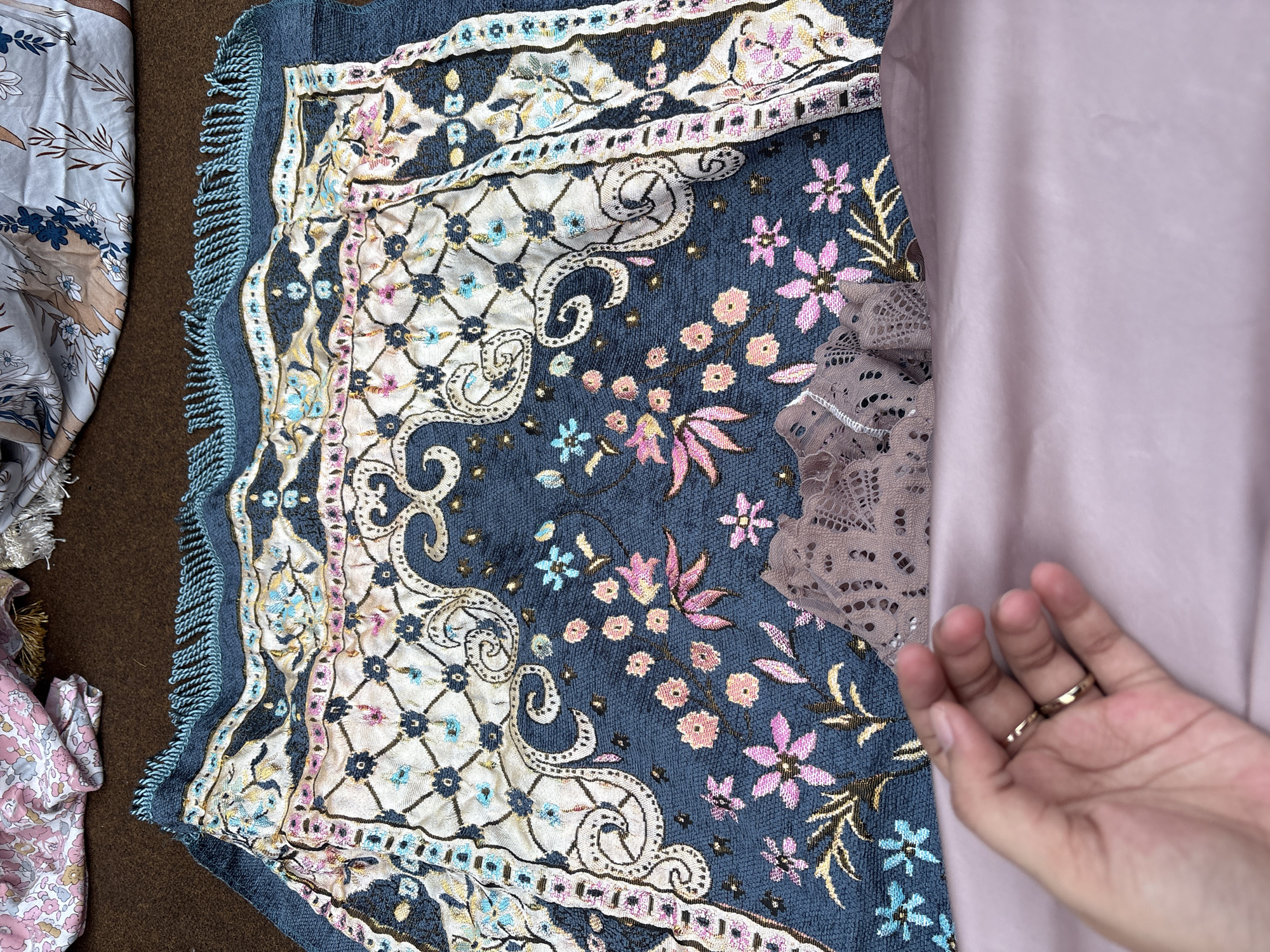 The image presents a top-down view of a textile arrangement. The primary focus is a blue prayer rug adorned with floral and paisley patterns, framed with fringe. Adjacent to it are fabric pieces with floral prints, a pink cloth, and a piece of lace. The right side of the image captures a hand with gold rings. The location, Gadingrejo, Indonesia, provides a geographical context but is not visually evident within the frame. The scene does not depict any specific actions or interactions, primarily featuring static objects.
