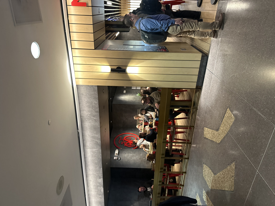 People stand in a line near a restaurant. The restaurant features a long counter with red stools where patrons are seated and eating. A large, stylized red logo is visible on the wall. Arrows are inlaid in the floor, pointing the way. The restaurant has a modern interior with light wood accents on the exterior and gray walls inside.
