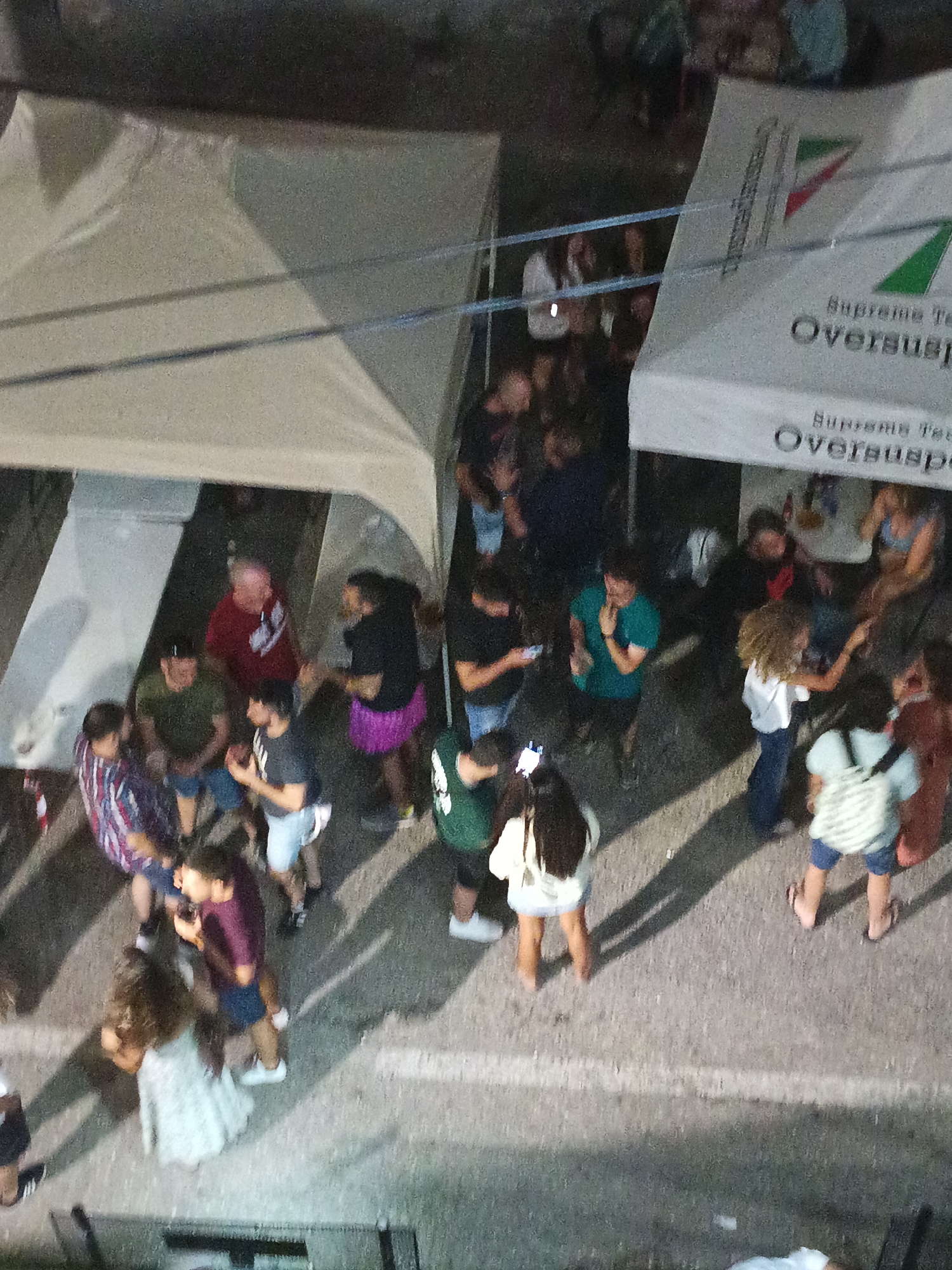 This aerial, slightly blurred image captures an outdoor gathering of people near tents. The environment appears to be an urban or semi-urban setting, possibly a street or plaza, indicated by the paved ground with white lines.
In the foreground, a group of individuals are visible, some in casual attire. One person in a white dress is standing, while another in a burgundy shirt and dark shorts stands next to them, holding a phone. To their left, a person wearing a striped shirt is interacting with another individual. Further back, a group of people are standing and talking.
Two large white tents are prominent in the scene. One tent on the left is open, and the other on the right displays a logo with Italian colors and text that includes "Supreme Tex" and "Oversuspen". There are also some bottles on a table near the right tent, suggesting food or drink service.
The overall impression is of a social event or a small festival. The lighting suggests it might be late afternoon or early evening. The specific location of Fuentesaúco De Fuentidueña, Spain, suggests this event is taking place in a public space within that municipality.