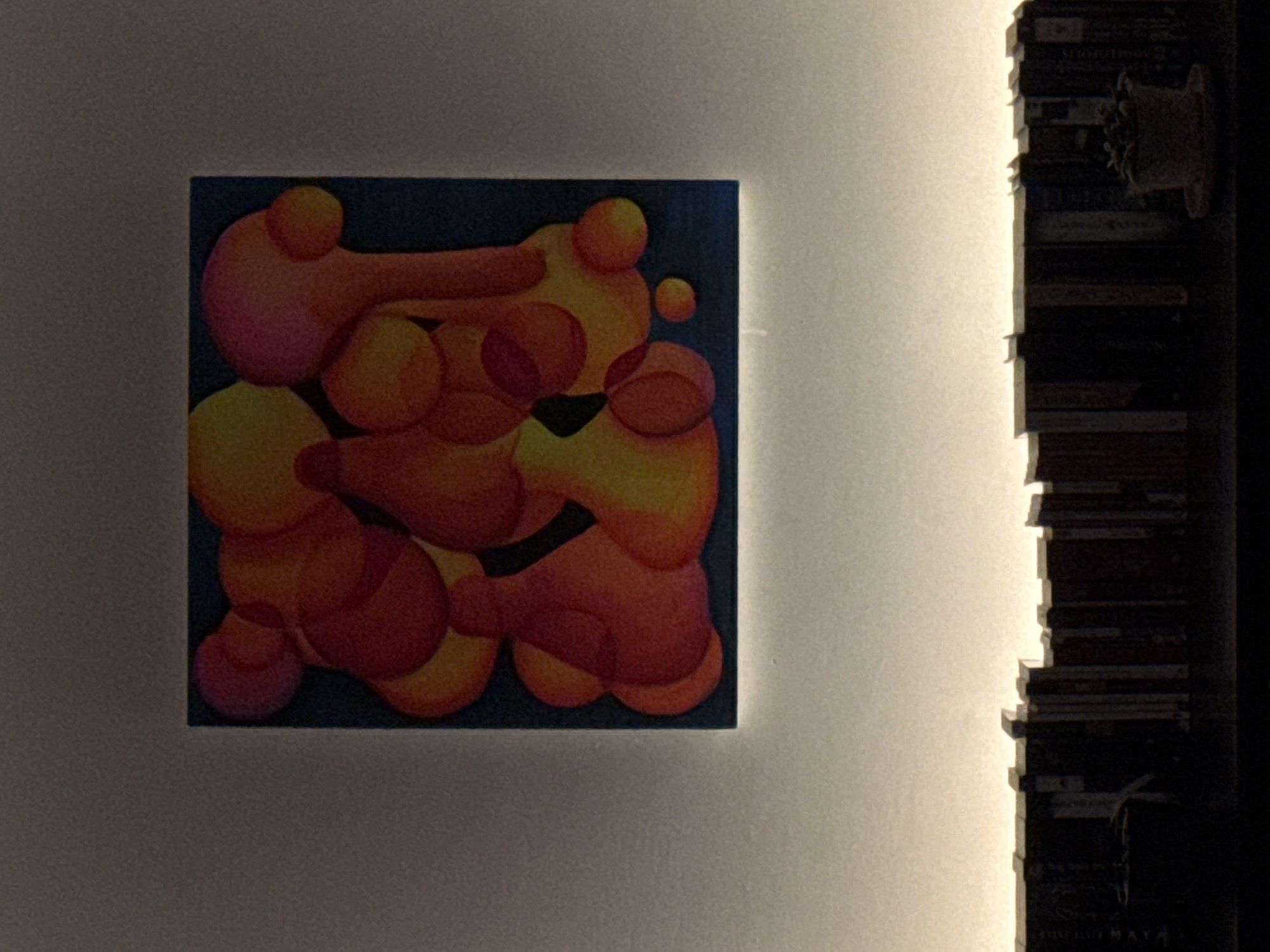 An abstract painting hangs on a white wall, bathed in warm light, possibly from a concealed source. The painting features a blue background with overlapping orange and red bulbous shapes. Adjacent to the wall is a dark bookcase, its shelves stacked with books. The location is indiscernible from the provided image.