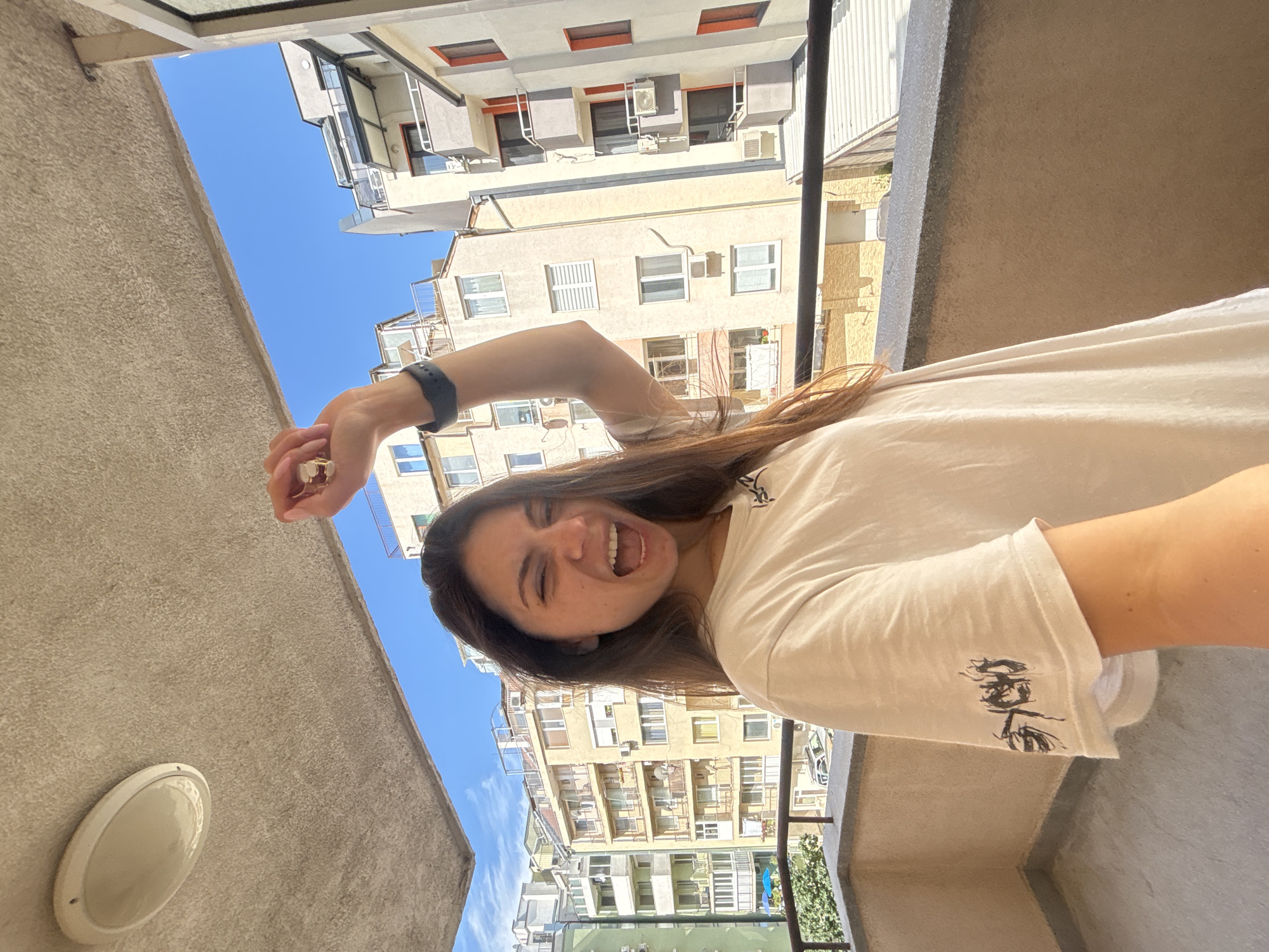 A young woman with long brown hair is standing on a balcony in Varna, Bulgaria, holding up her right hand and making a fist. She is wearing a white t-shirt with a black graphic design on the sleeve and a black smartwatch on her left wrist. She is winking and has her mouth open as if cheering or shouting. Behind her, there are several apartment buildings and a bright blue sky. The balcony has a concrete railing and ceiling.