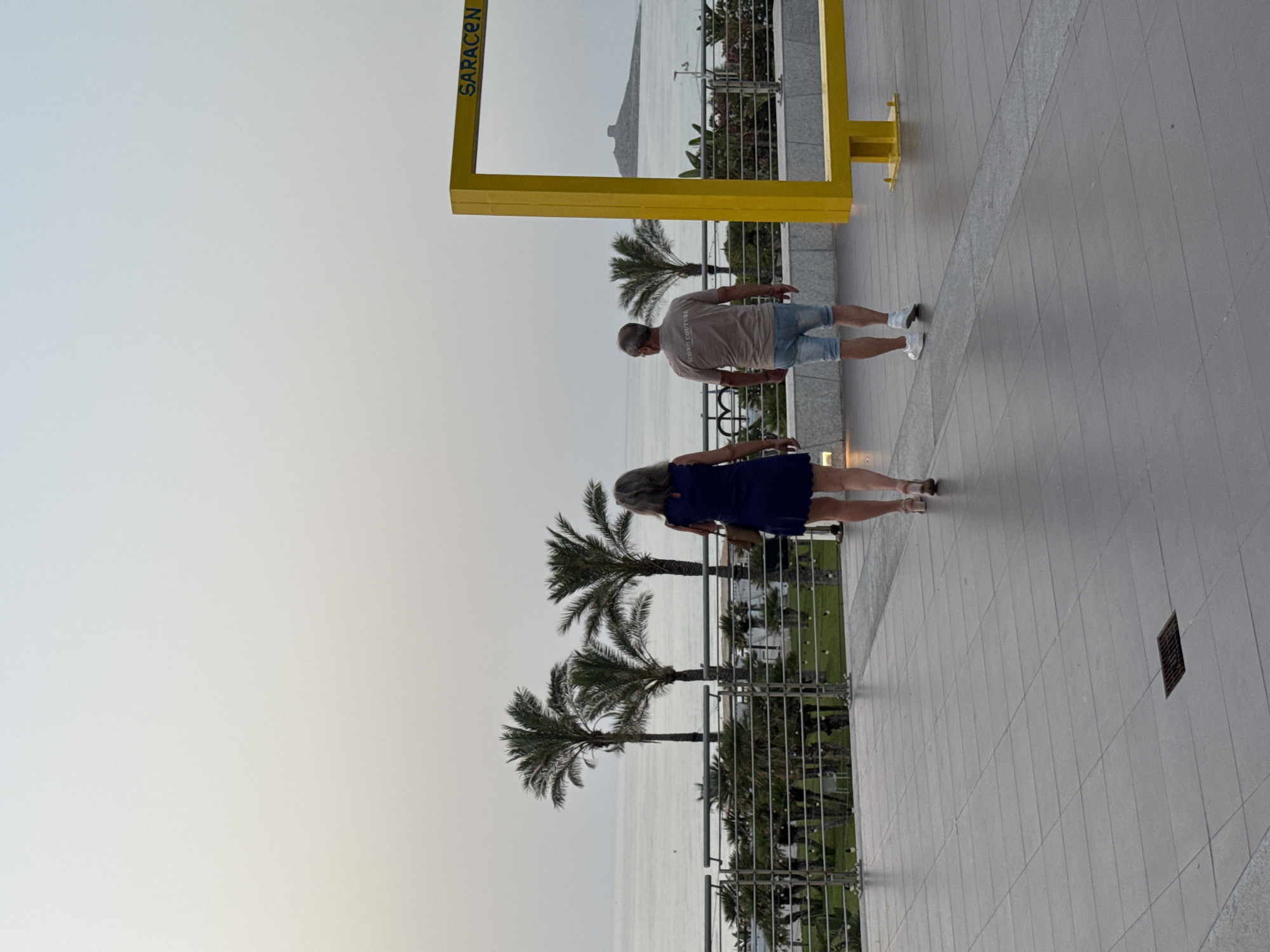 Two people, a woman in a dark blue dress and a man in a tan t-shirt and denim shorts, are walking away from the camera on a tiled walkway. They are facing the sea, with palm trees and a railing in the foreground. In the background, the sea stretches to the horizon under a hazy sky. To the right of the frame, a bright yellow, rectangular structure is visible, with the word "SARACEN" written in yellow on a brown background. Further in the distance, a small island with a tower on it is visible in the sea. The overall scene suggests a coastal resort or promenade.