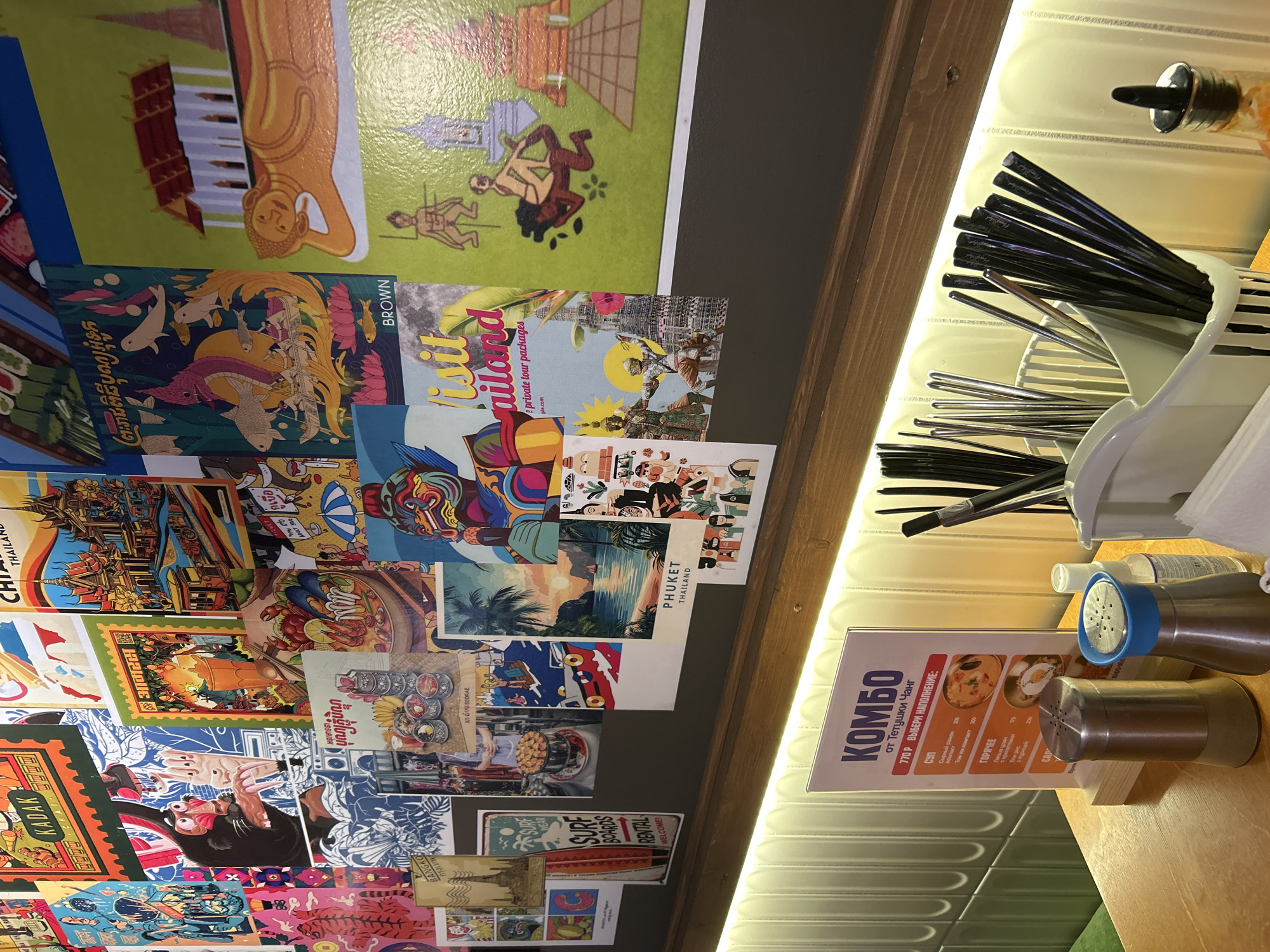The image captures a scene within an eatery, possibly in Izhevsk, Russia, given the presence of Russian text on the menu. A section of the wall is decorated with postcards or posters featuring images of Thailand, indicating a theme or type of cuisine offered by the establishment. In the foreground, there is a table surface on which a stand displaying a menu with "KOMBO" in bold (Russian for Combo), and a price of 770 Rubles, suggests a special offer. The menu is in Russian with dishes like "Soup: Cheese Ramen and Tom Yam Compact" and "Hot: Udon noodles with shrimp and Wok with Chicken". Condiment dispensers and a container with chopsticks and other utensils are also present on the table. The background shows a textured wall with a light fixture. The image appears to have been taken from an angle looking up at the wall decorations and table setting.