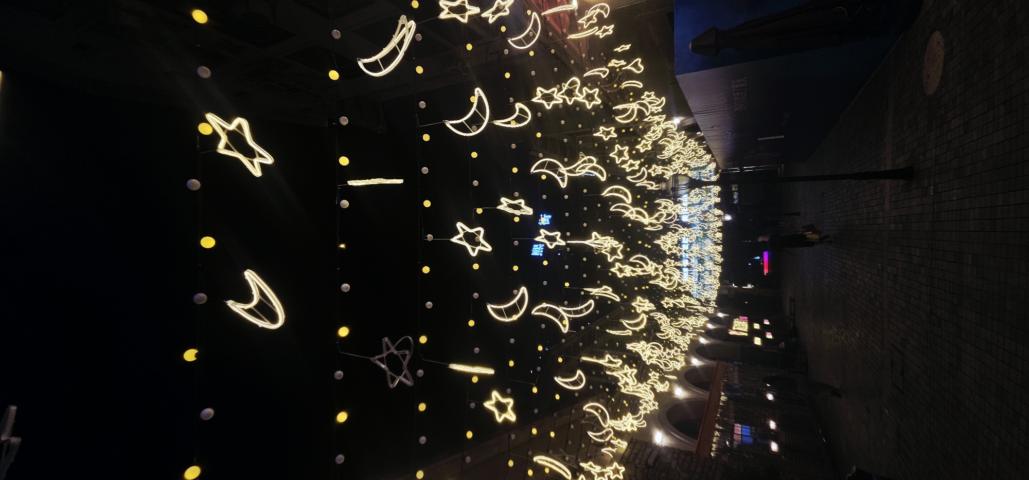 The image shows a night scene in an outdoor pedestrian area, possibly a shopping street or an entertainment district, with numerous string lights shaped like stars and crescent moons suspended overhead. The lights are a bright yellow and are illuminated against the dark night sky. There are also smaller, round yellow lights and some vertical light elements interspersed. The ground is paved with brick or stone, and there are architectural elements visible on the left, appearing to be archways of a building with interior lights. A lamppost stands on the right side of the pathway. Several individuals are walking in the distance along the paved path, appearing as silhouettes against the ambient light. The setting suggests a festive or decorative atmosphere, likely in an urban environment. The location is identified as the Chongqing High-tech Zone, China.