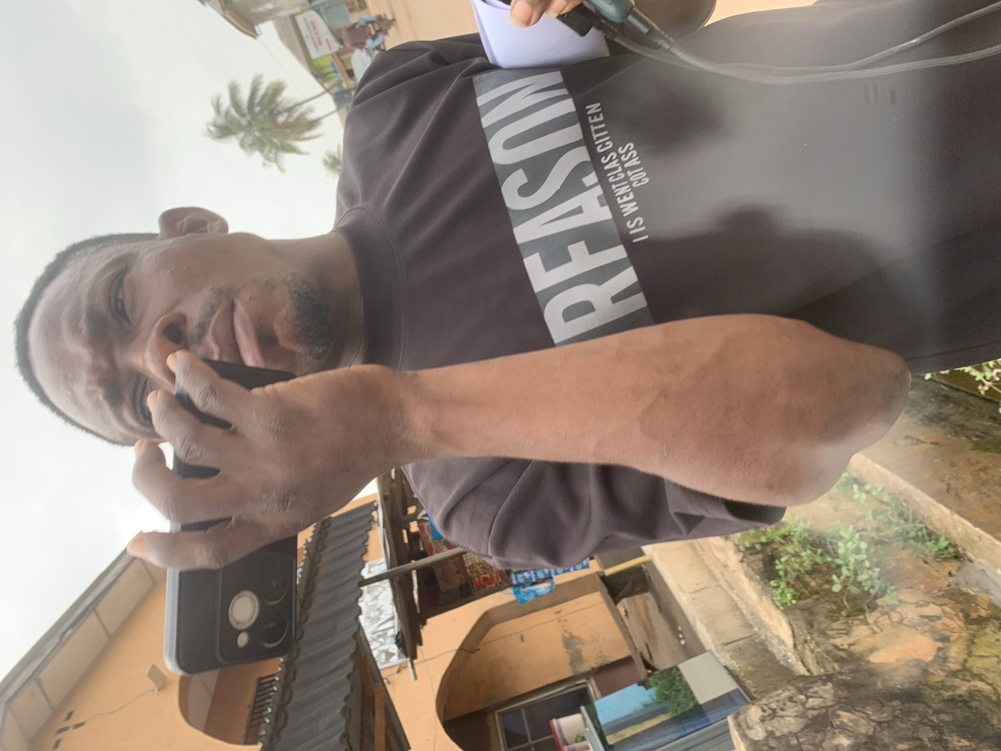 A close-up, head-and-shoulders, low-angle shot captures a Black man from the chest up, holding a smartphone to his ear, as if engaged in a phone call. He is outdoors on a cloudy day, with a muted color palette dominated by grays and earthy tones. The man is wearing a dark gray t-shirt with white lettering that reads "REASON" and below that, "I IS WENTCLAS CITTEN COT ASS." His skin is a medium-dark tone, and he has short hair and a goatee.
The background reveals an urban environment that suggests the location is Shagamu, Nigeria. To the left, an orange building with a dark roof and an arched doorway is visible. In the mid-ground, there's a makeshift stall with items displayed on shelves, hinting at a market or roadside vendor setting. A slight sheen on the t-shirt suggests it might be slightly wet or under a reflective surface. To the right, a paved road is visible, with a few indistinct structures and foliage, including a palm tree, further indicating a tropical or subtropical setting. The overall impression is that of an everyday street scene in a bustling town.